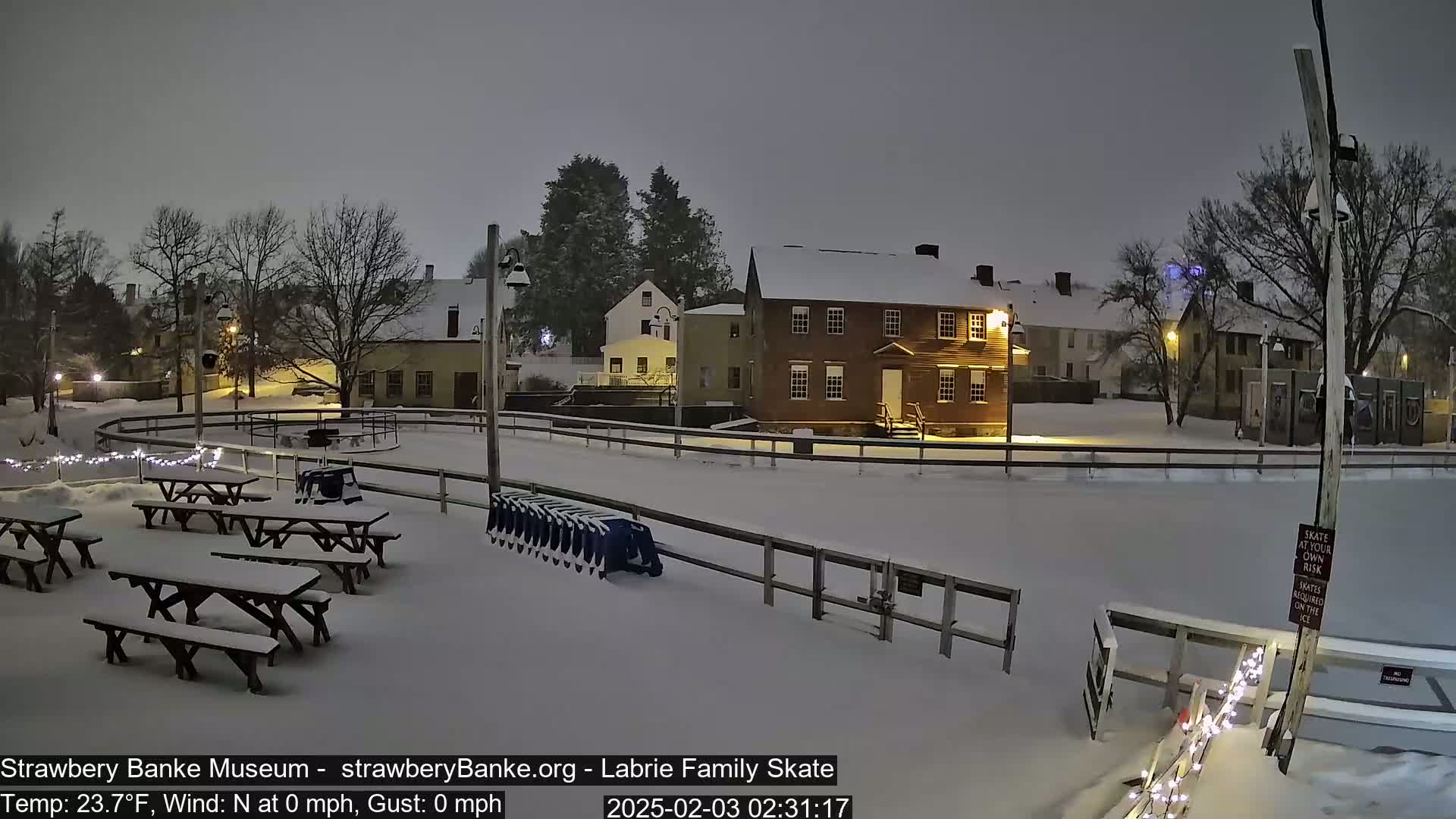 A snow-covered outdoor ice rink with picnic tables and a building is illuminated at night during a light snowfall.