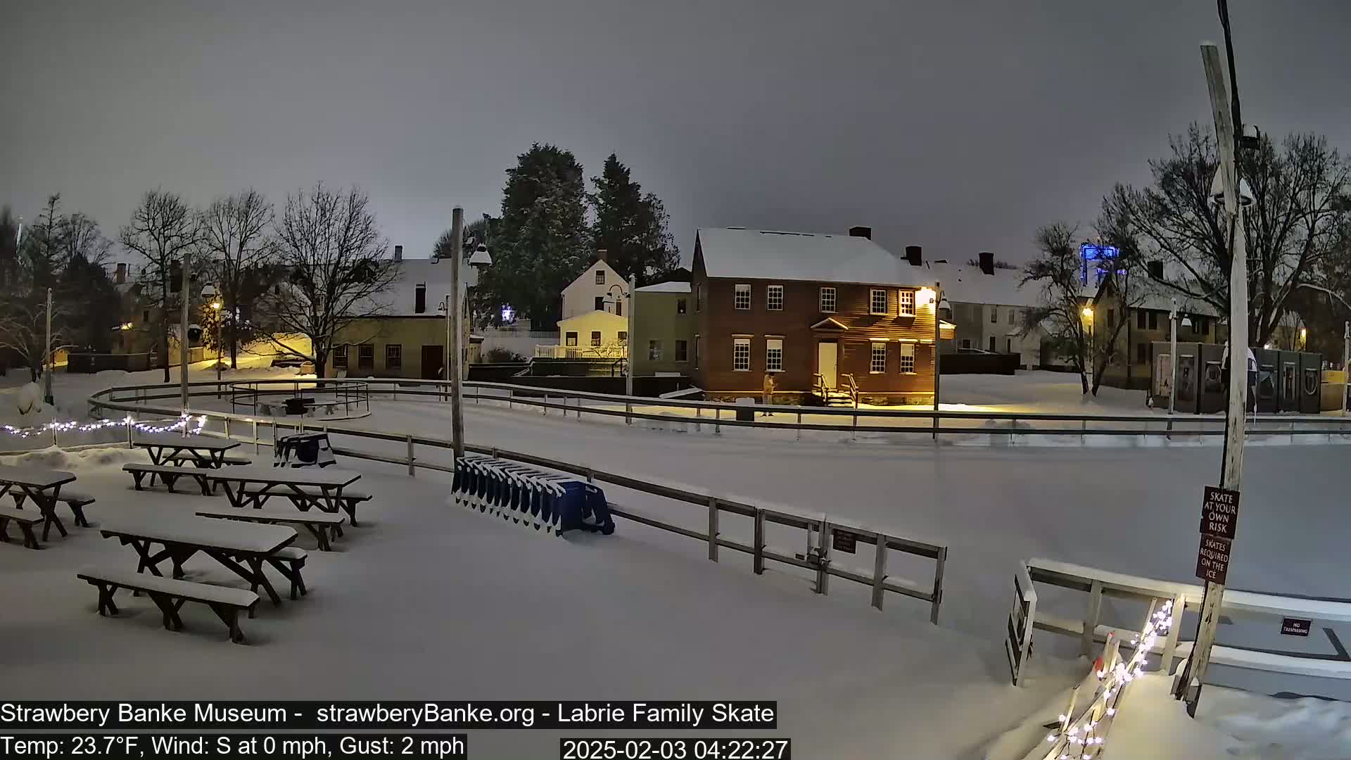 A snow-covered outdoor ice skating rink is surrounded by snow-covered picnic tables, buildings, and trees under a dark, overcast sky.
