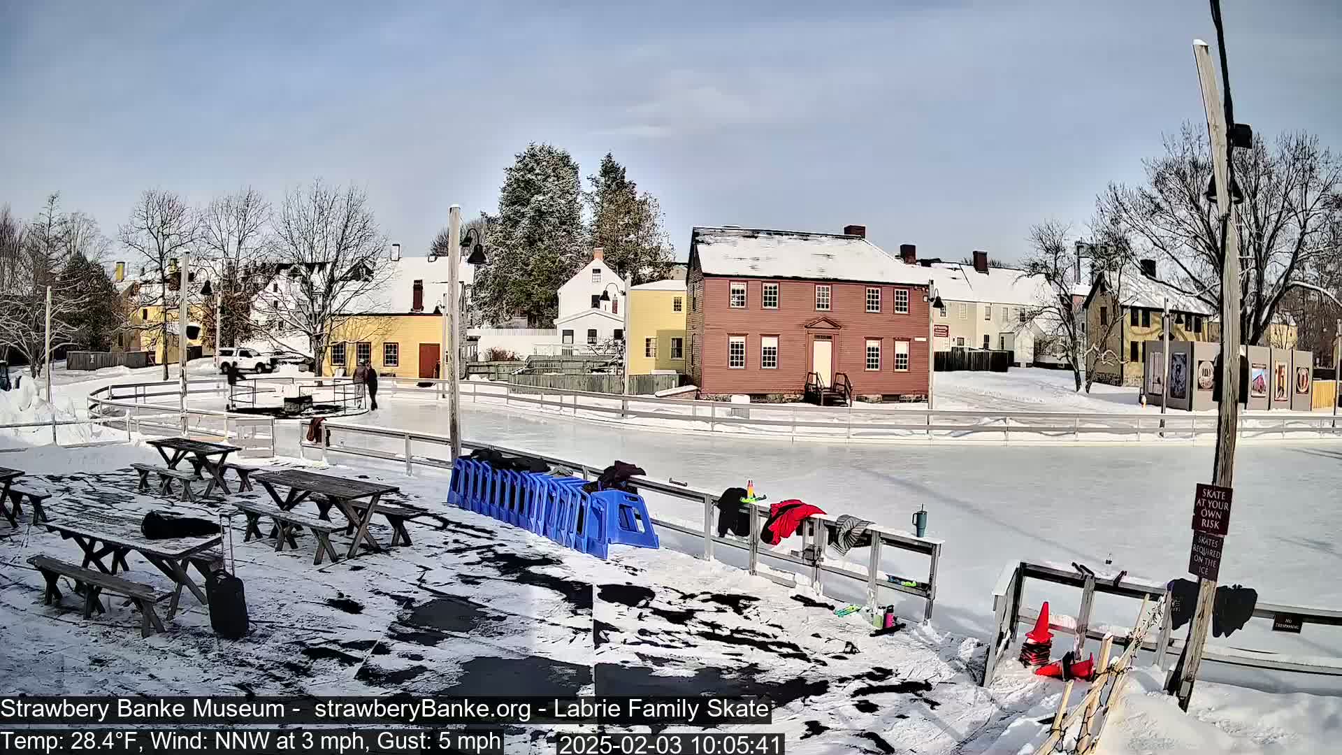 A snow-covered outdoor ice rink is situated next to several historic buildings under a sunny sky, with picnic tables and skate-related equipment nearby.