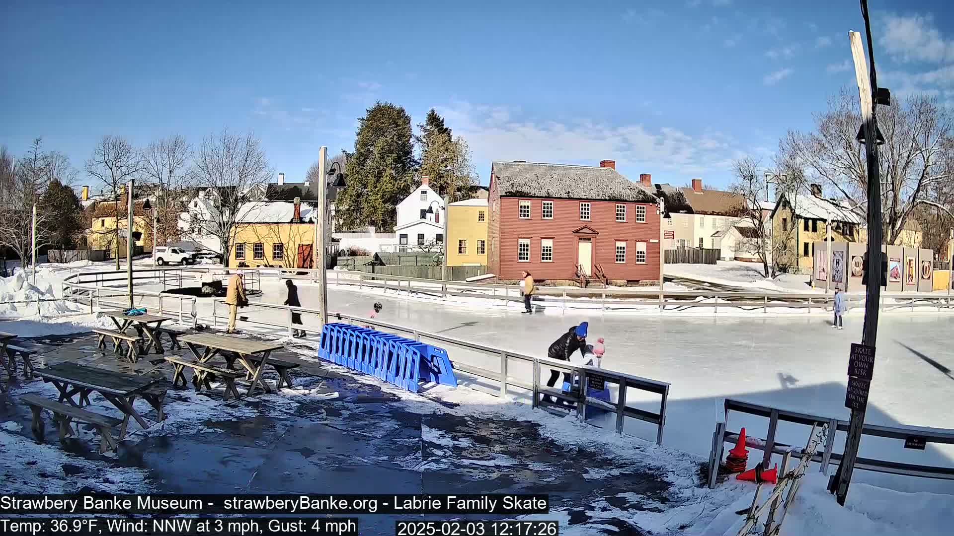 A sunny outdoor ice-skating rink is surrounded by snow-covered ground and historic-looking buildings, with several people ice skating and others standing near picnic tables.
