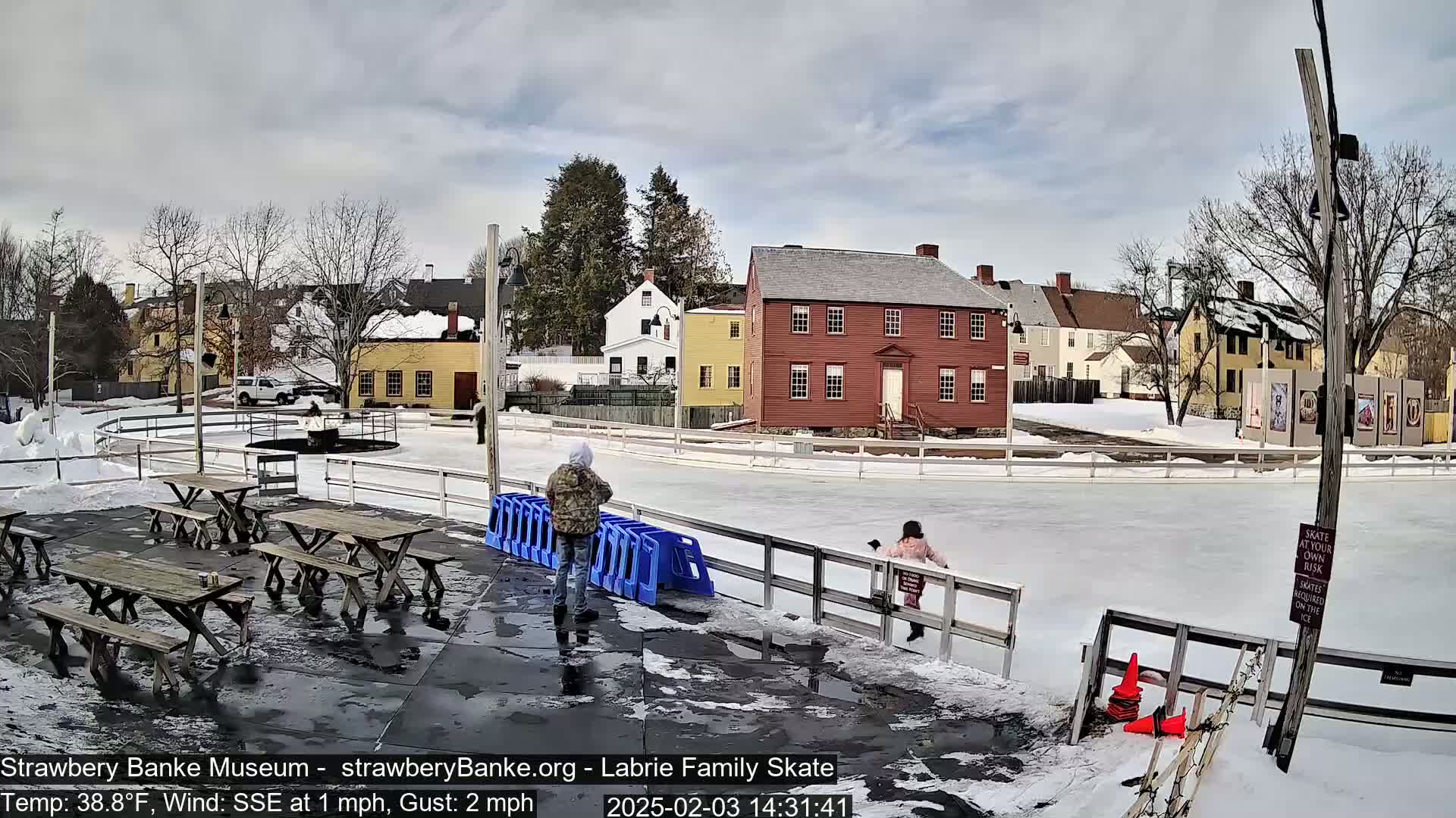 An outdoor ice skating rink, partially melted in places, is surrounded by historic-looking buildings under a mostly cloudy sky.