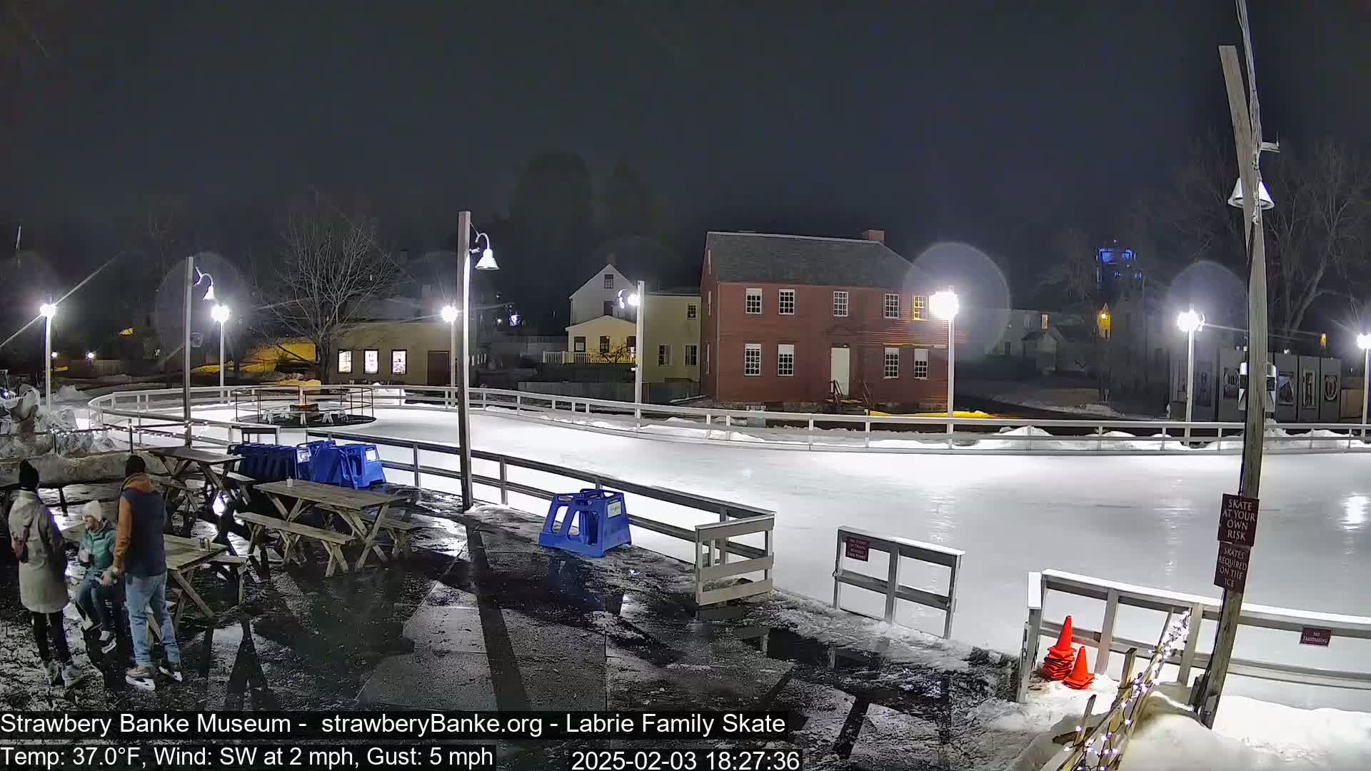An outdoor ice rink is lit at night under partly cloudy skies, with a few people standing near picnic tables beside it and a red brick building in the background.