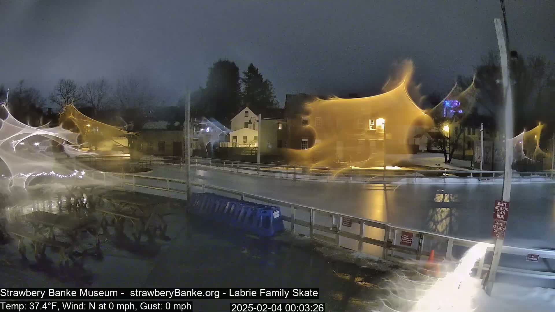 A nighttime view of an outdoor ice rink with some snow on the ground, illuminated by nearby buildings and streetlights, with what appears to be blowing snow or ice crystals creating streaks of light in the image.