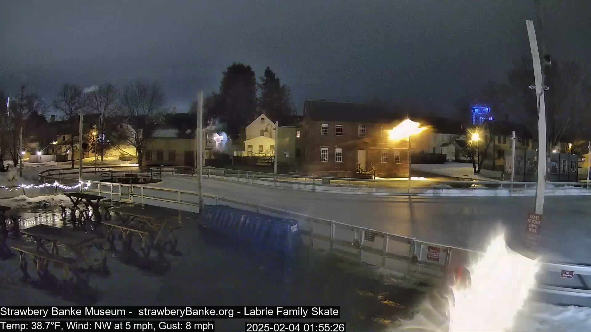 A nighttime view of an outdoor ice rink near several buildings, under a cloudy sky with a northwest wind.