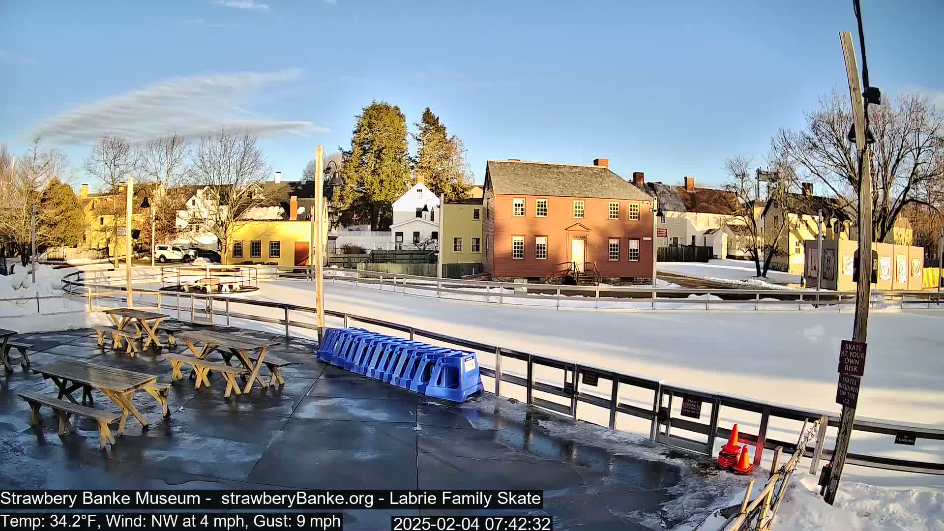 A sunny, cold day reveals an outdoor ice skating rink surrounded by historical buildings and wooden picnic tables.