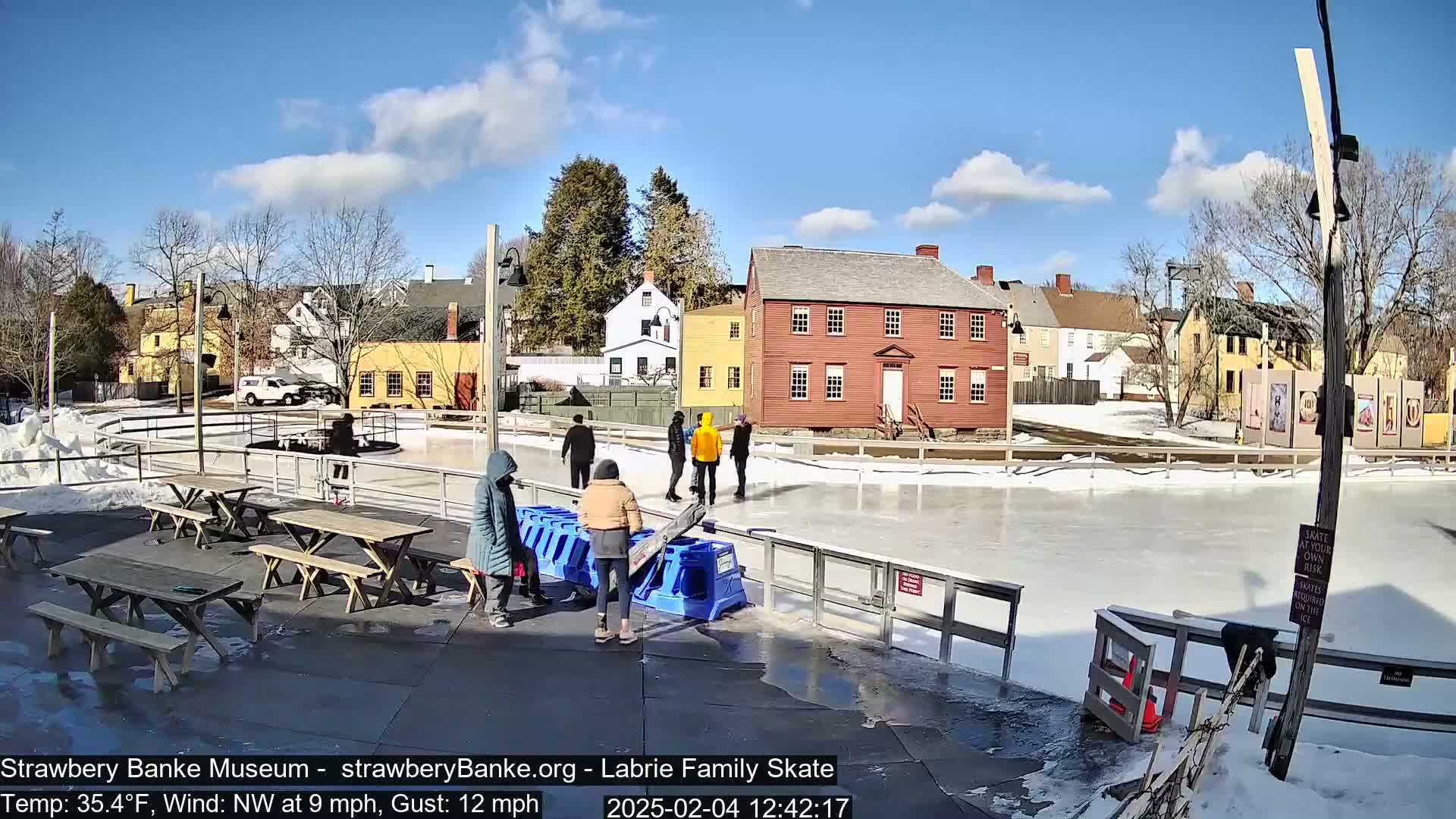 Several people are ice skating on an outdoor rink in front of historic buildings on a sunny day with a northwest wind.