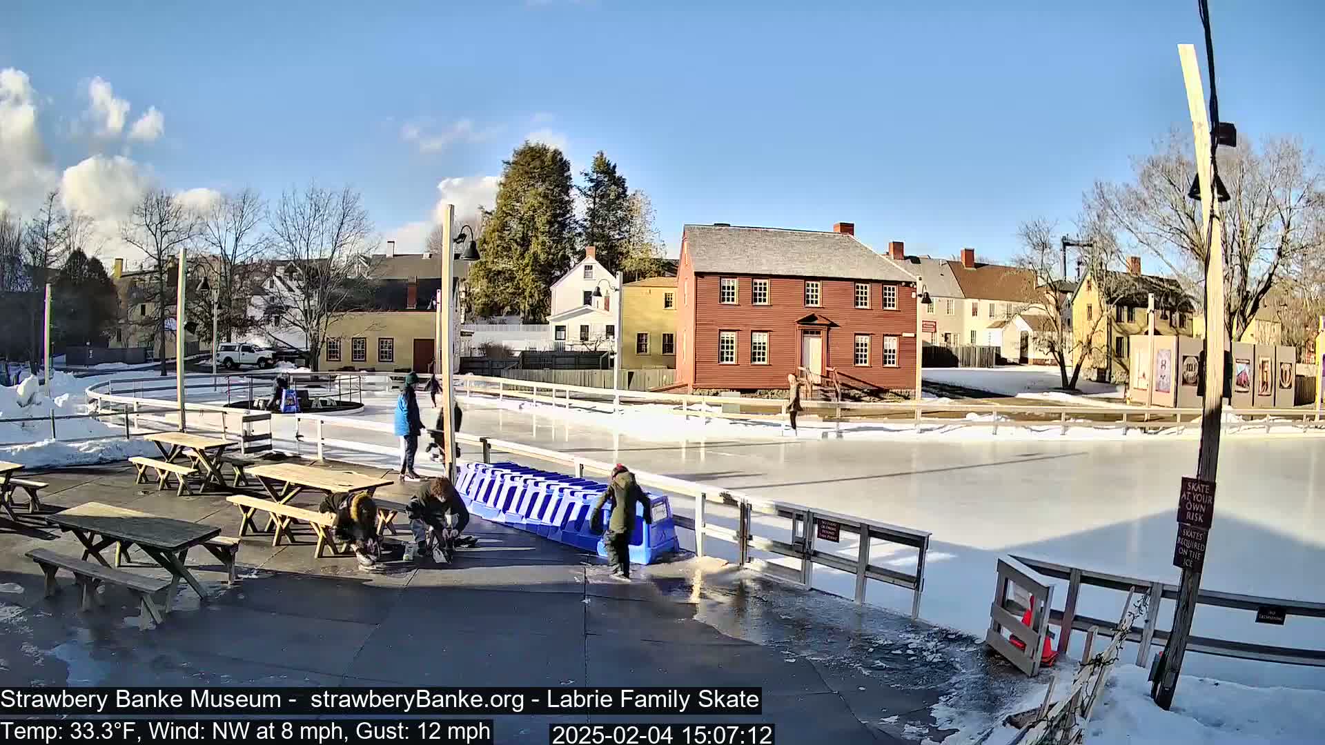 A sunny outdoor ice skating rink, surrounded by historical buildings and picnic tables, has several people skating and some people near the edge.