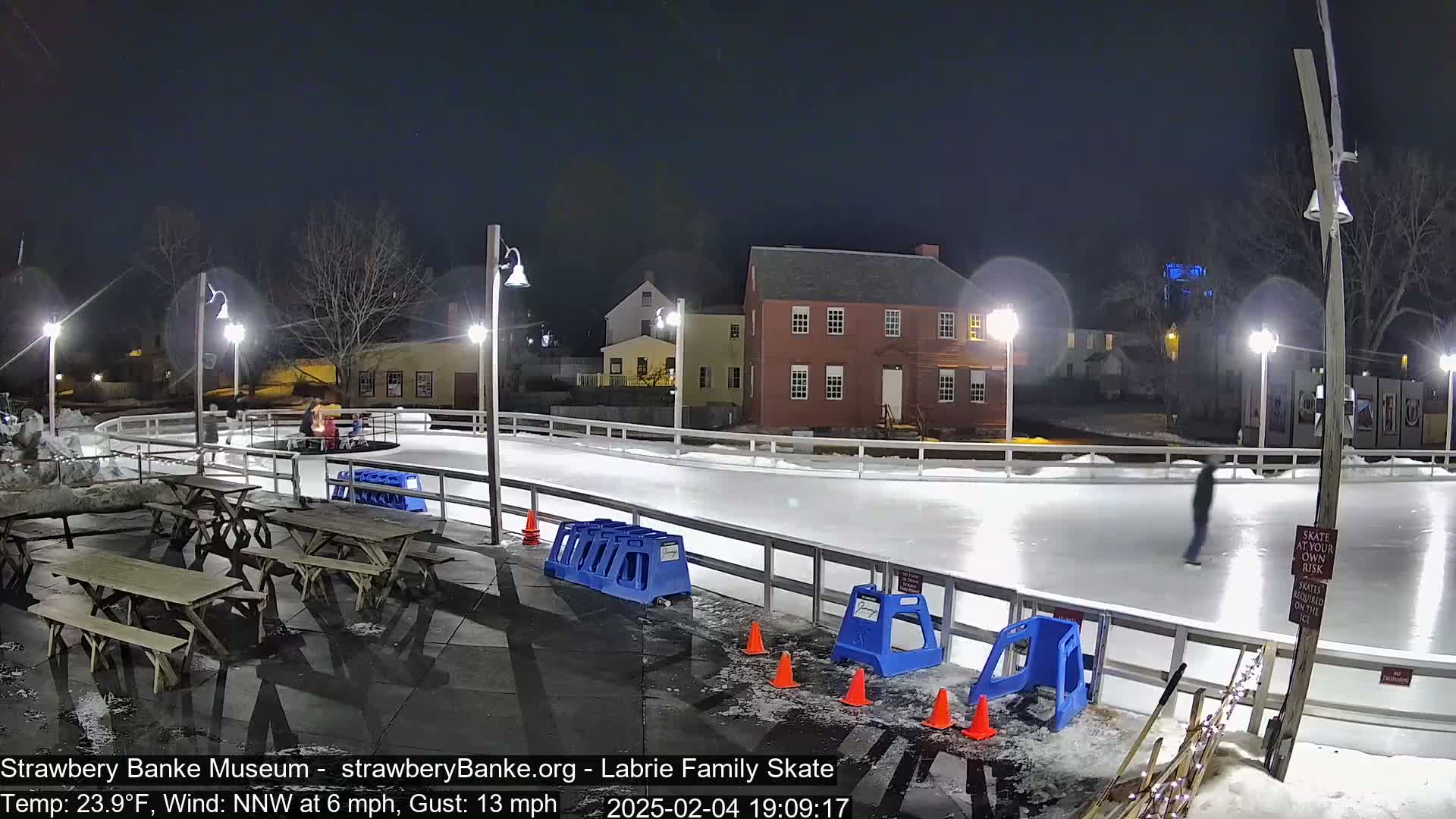 An outdoor ice skating rink is illuminated at night by several lamps, with a few people skating and others sitting at picnic tables nearby.
