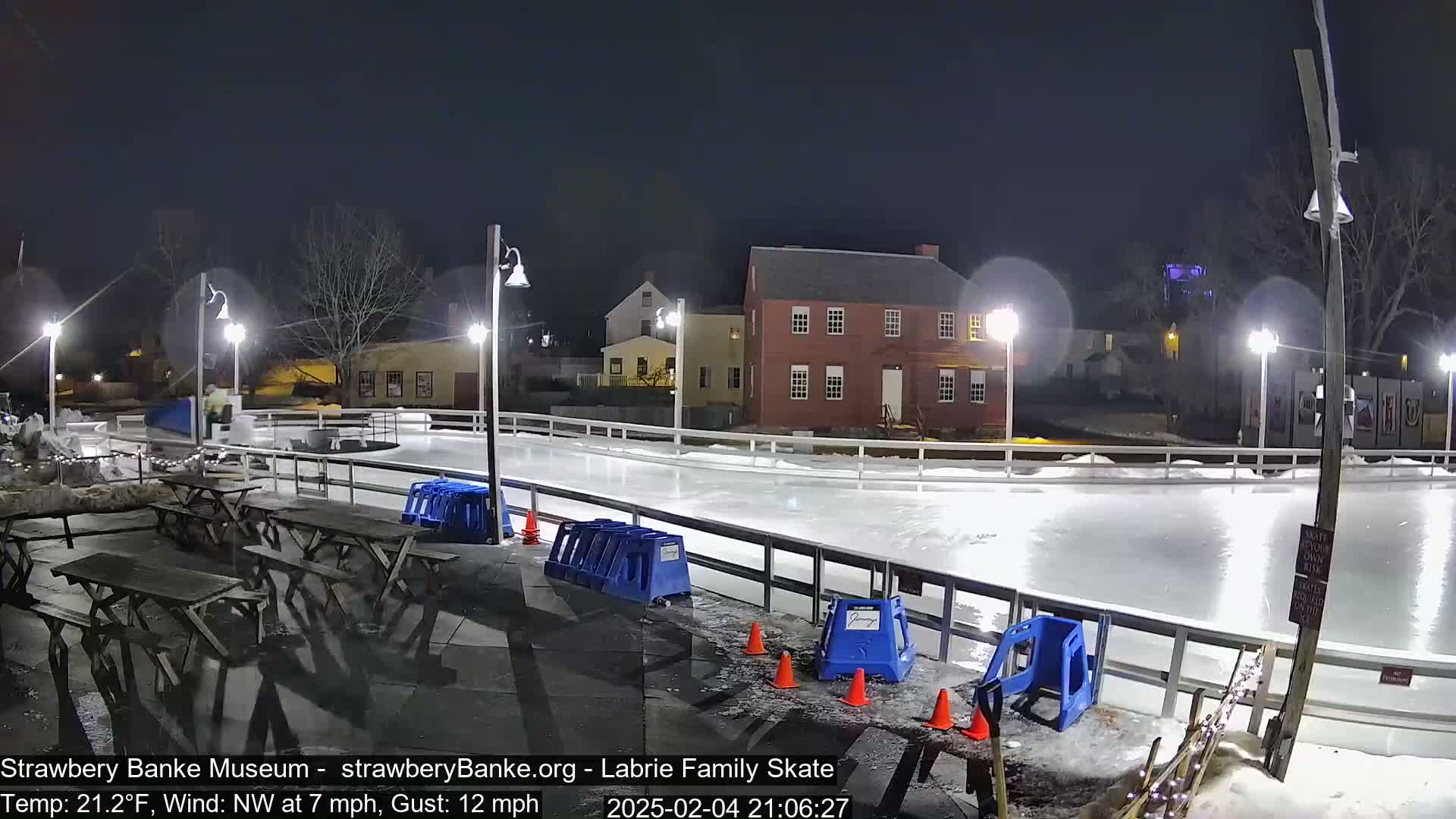 An outdoor ice skating rink is illuminated at night under a clear, dark sky, with picnic tables and blue plastic seating nearby.