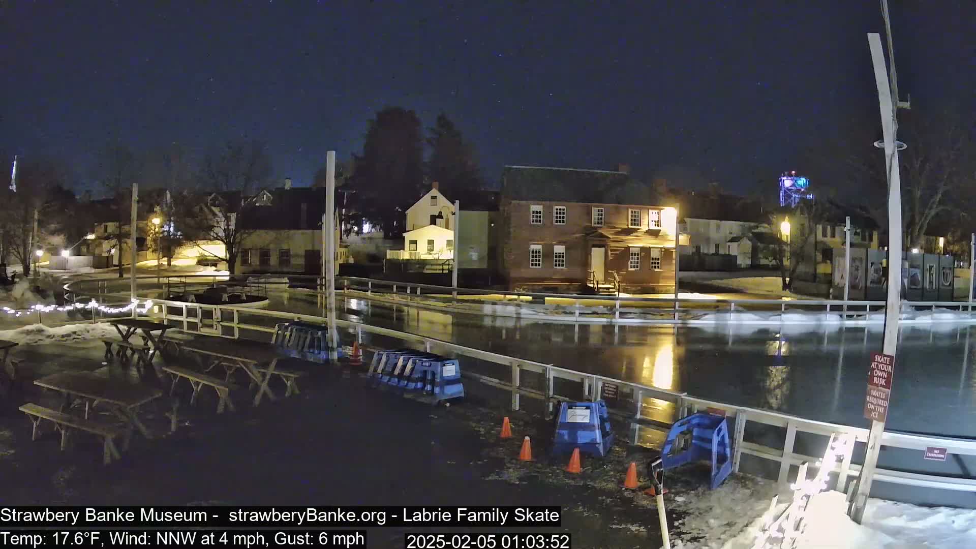 Under a starry night sky, a partially flooded outdoor ice skating rink is surrounded by buildings and picnic tables.
