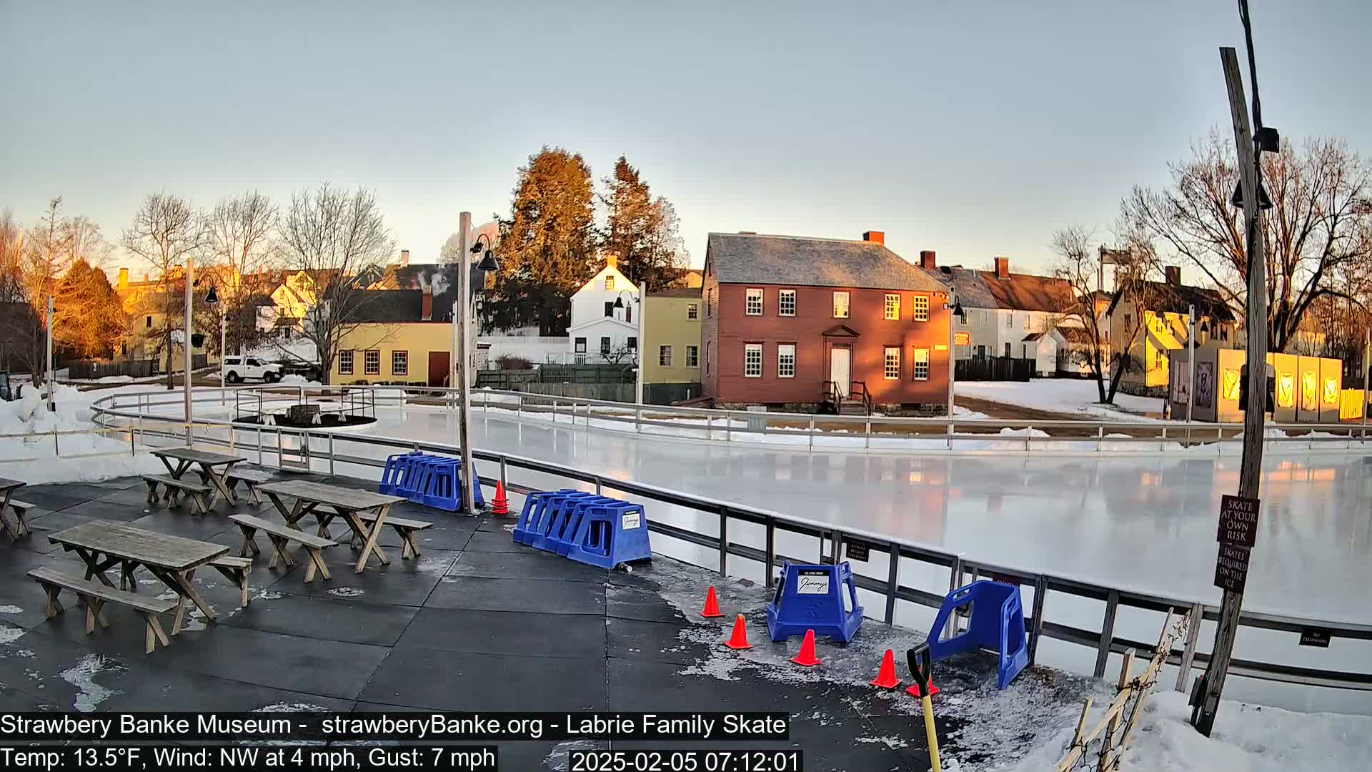 An outdoor ice rink sits in front of a row of historic buildings during a cold, sunny morning with a northwest wind.