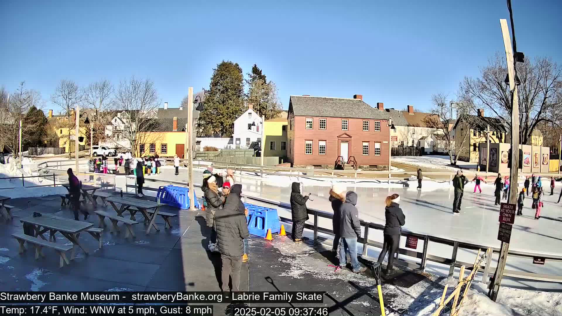 On a sunny, cold day with a WNW wind, people ice skate on an outdoor rink near several historic buildings.