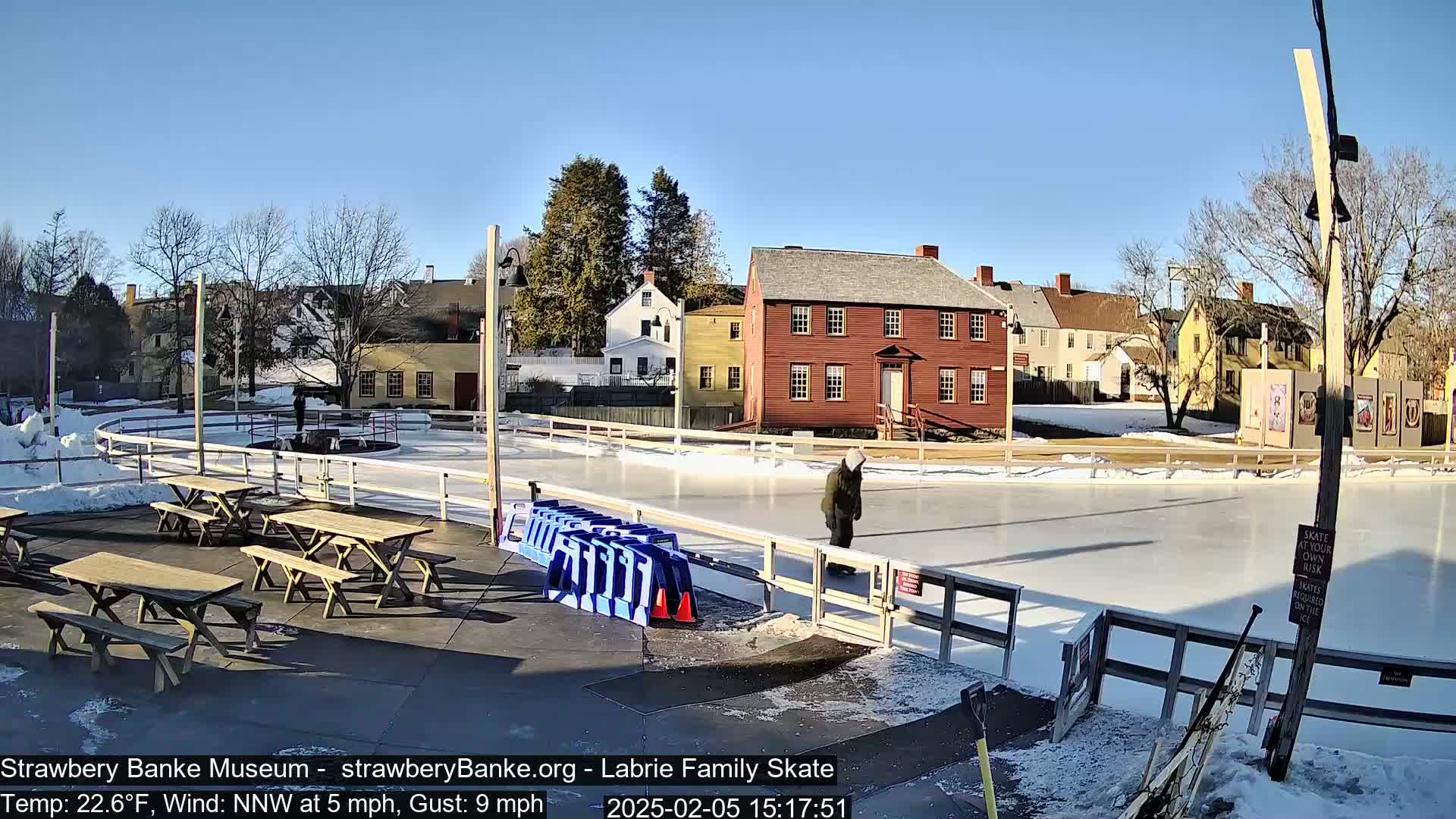A person ice skates on an outdoor rink surrounded by historic buildings on a sunny day with a 22.6°F temperature and a 5 mph northwest wind gusting to 9 mph.