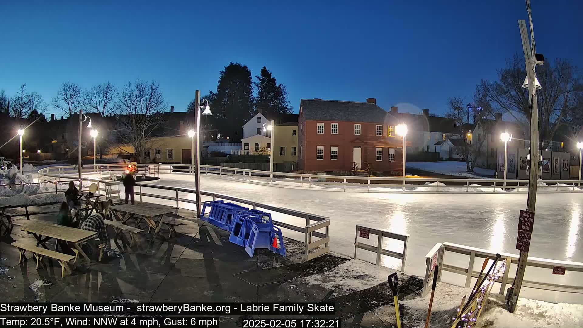 An outdoor ice skating rink is illuminated at night under a twilight sky, with people gathered near picnic tables beside it.