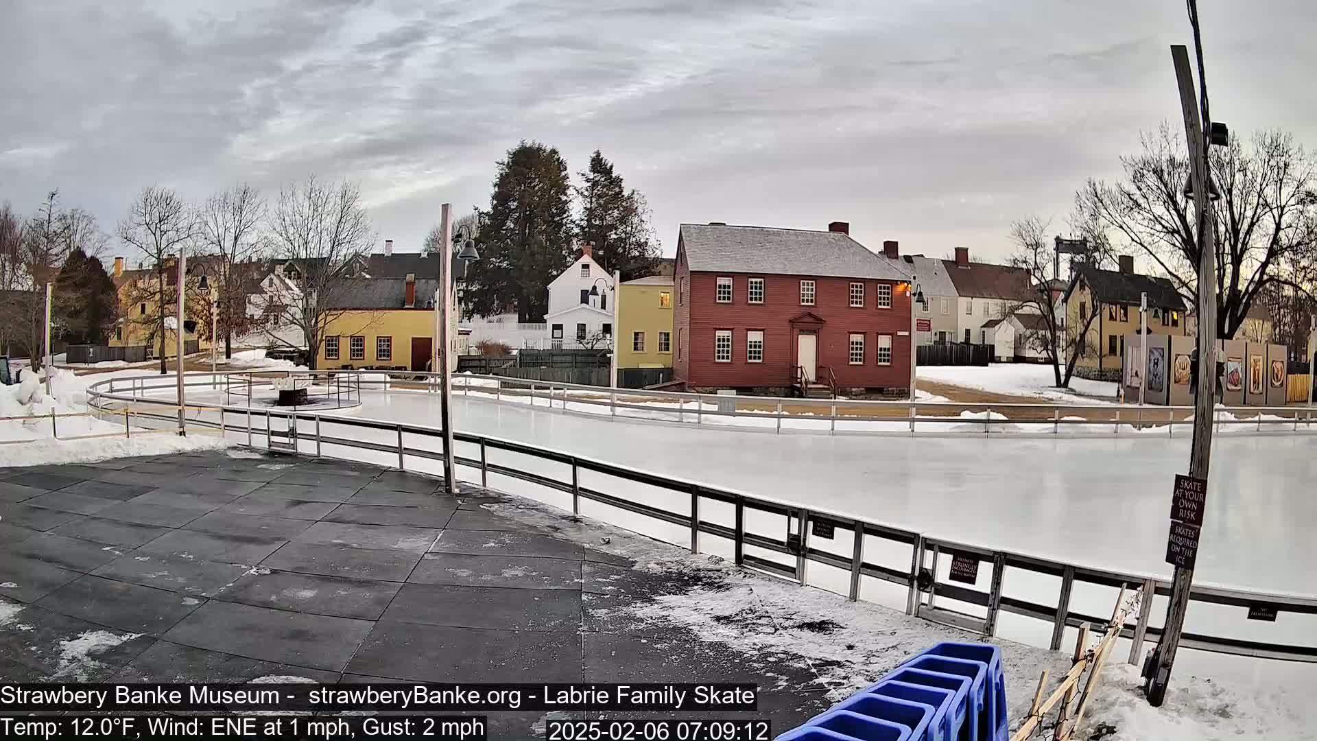 An outdoor ice skating rink, partially covered in snow, is situated in front of a cluster of historic-looking buildings under a cloudy sky.