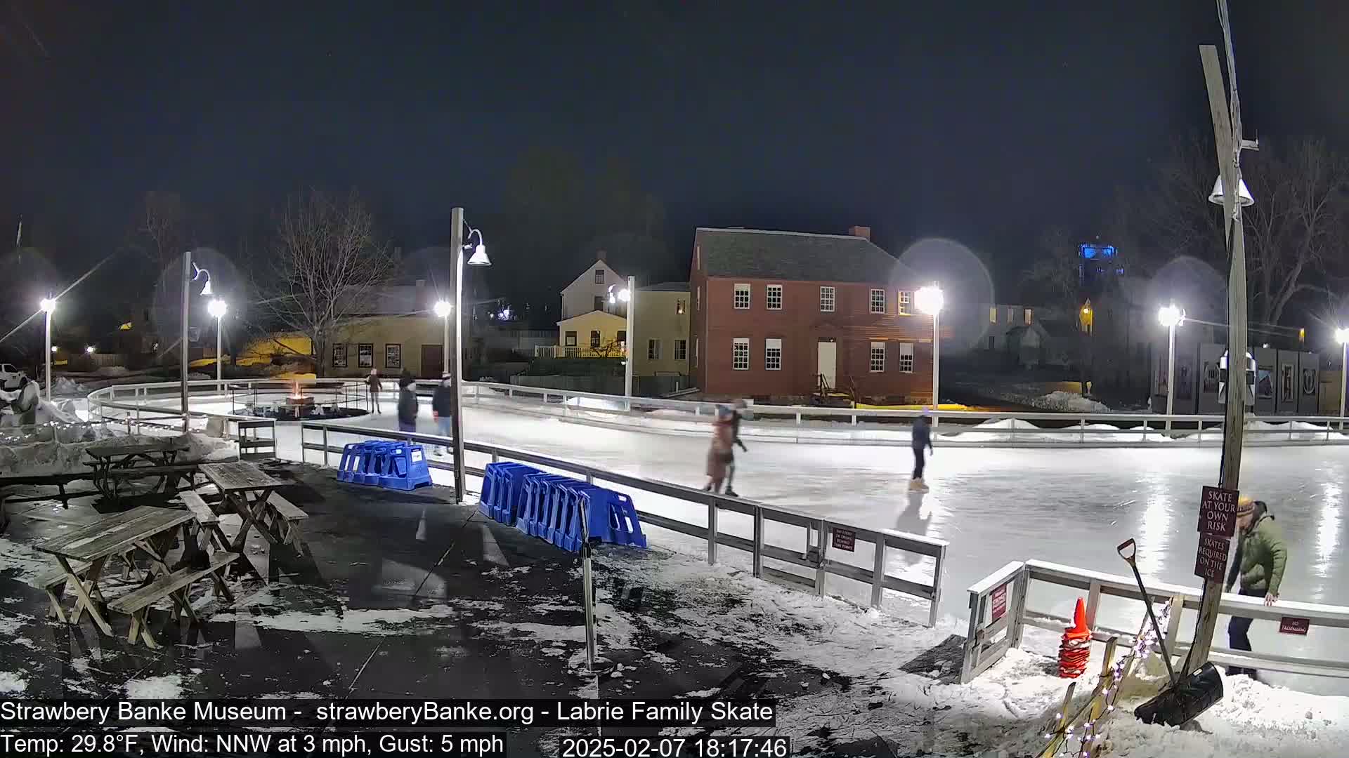 At night, under a clear sky, several people ice skate on an outdoor rink lit by lampposts, near some buildings and picnic tables.