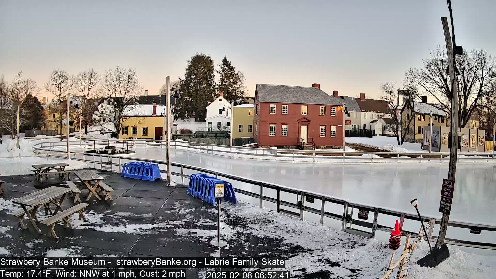 An outdoor ice skating rink, surrounded by picnic tables and historic buildings, on a partly cloudy evening with a temperature of 17.4°F and a light north-northwest wind.