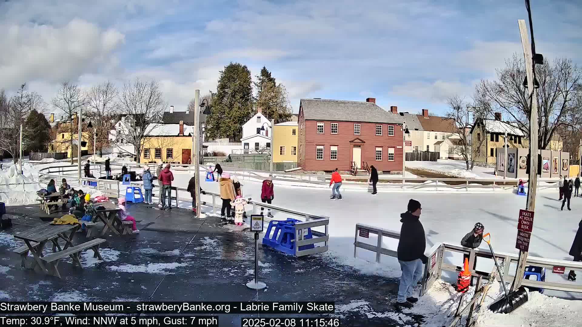 People ice skate on an outdoor rink surrounded by historic buildings under a mostly sunny sky.