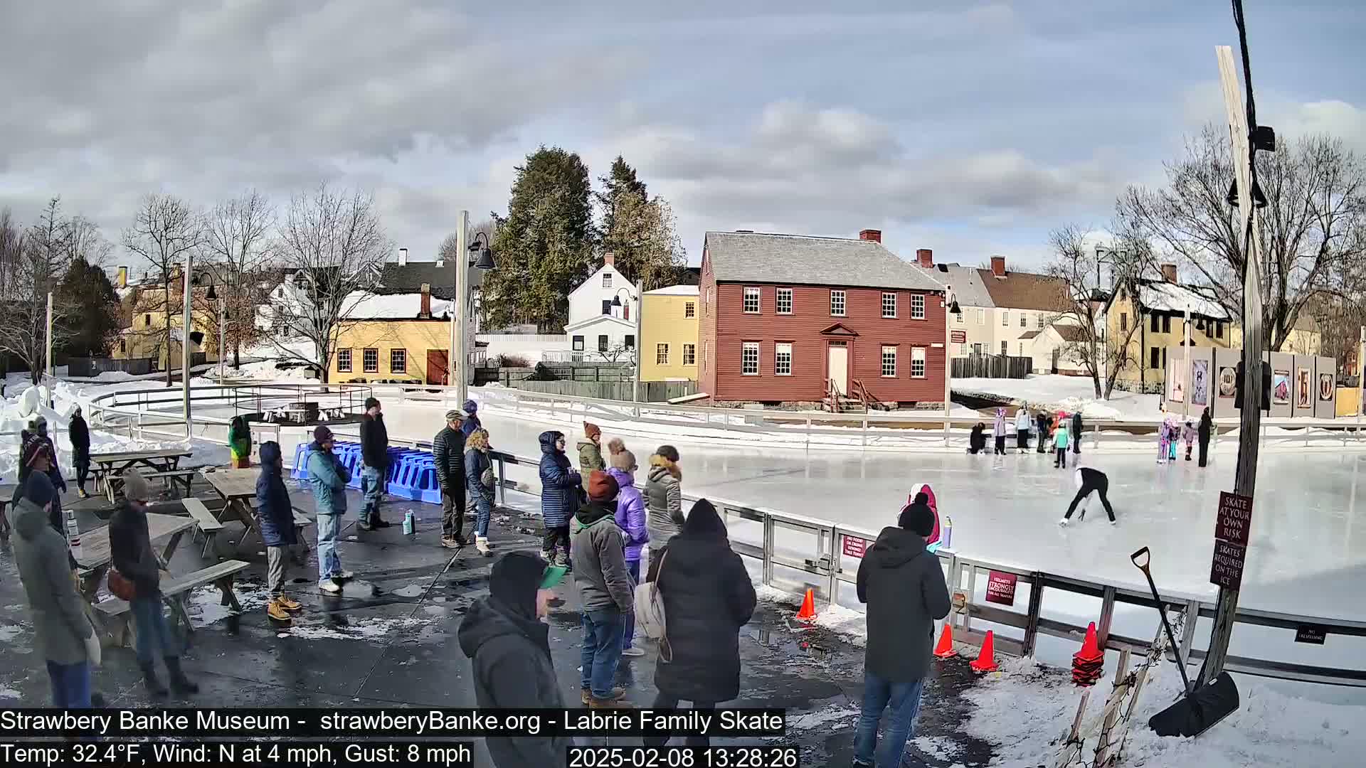 People stand around an outdoor ice skating rink on a partly sunny day with a light north wind.