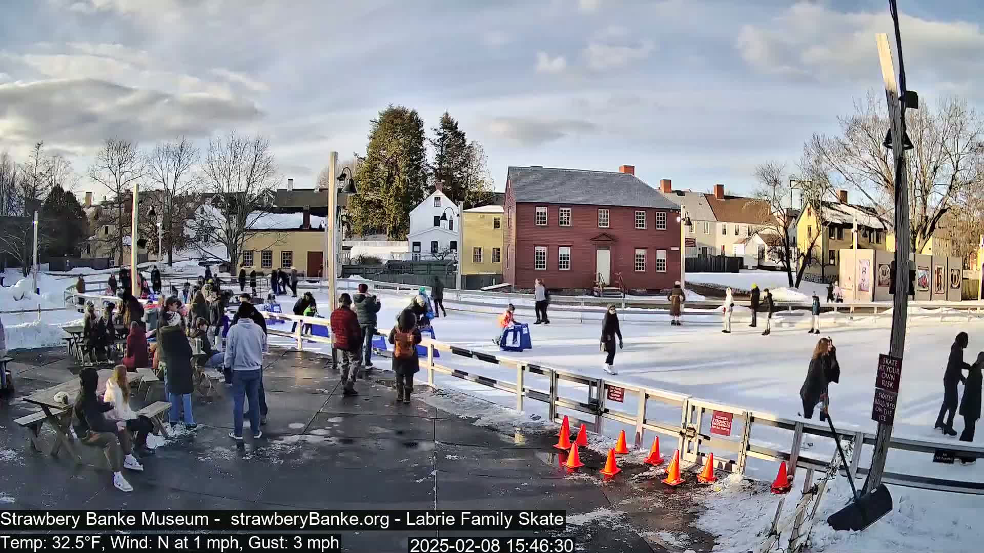 An outdoor ice-skating rink, surrounded by historic buildings, is populated with skaters and spectators on a partly sunny day with a light north wind.