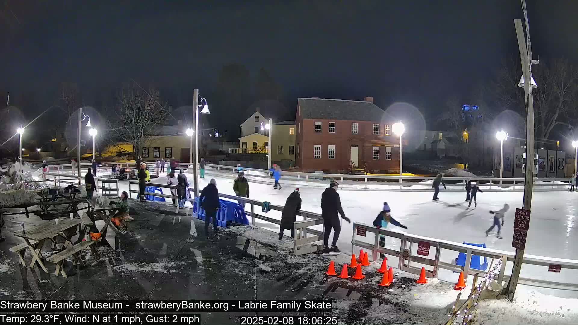 At night, under a dark sky, people ice skate on an outdoor rink illuminated by lights, near buildings and trees.