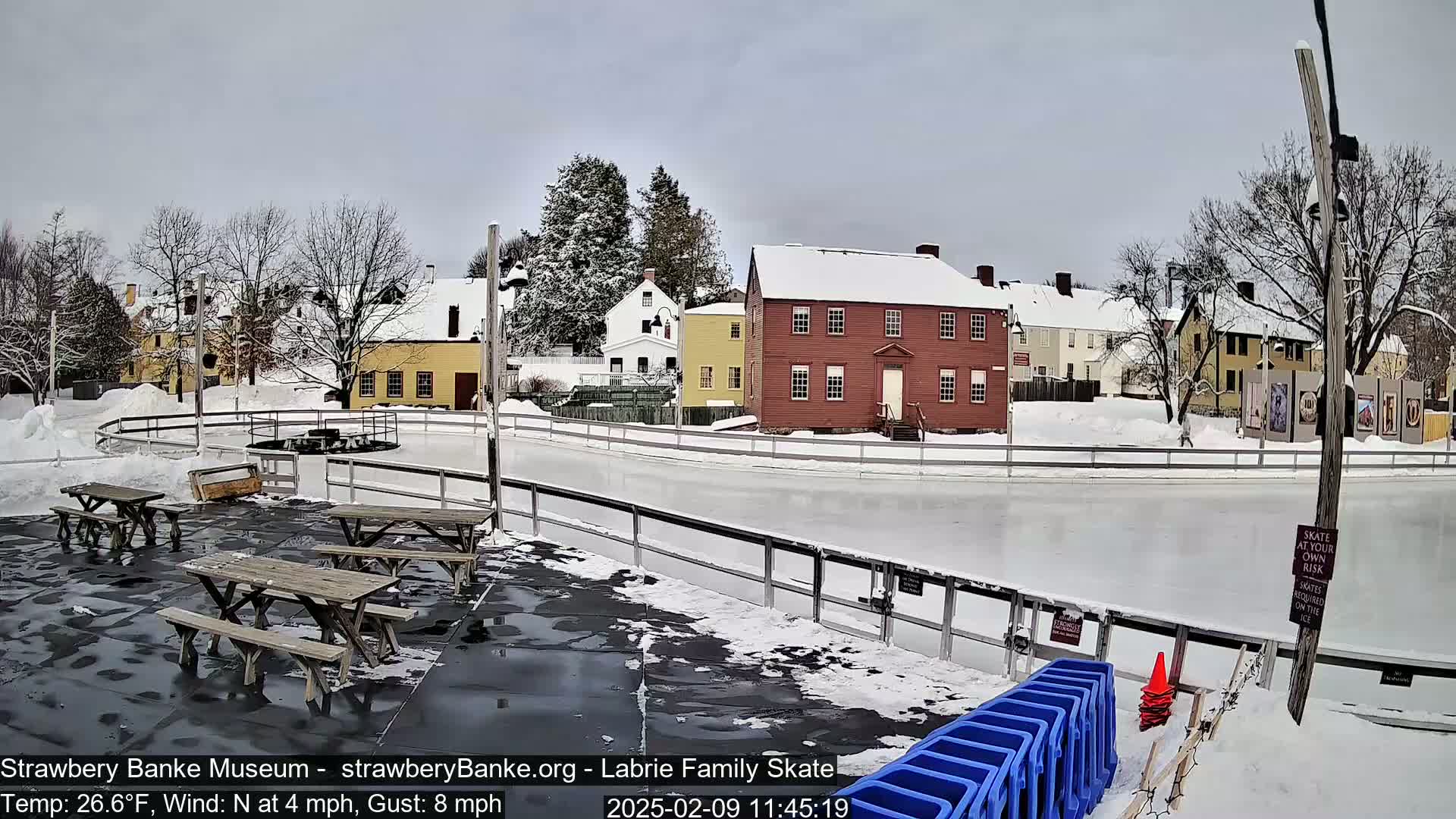 A snow-covered outdoor ice rink with picnic tables and several historic buildings in the background under a cloudy sky.