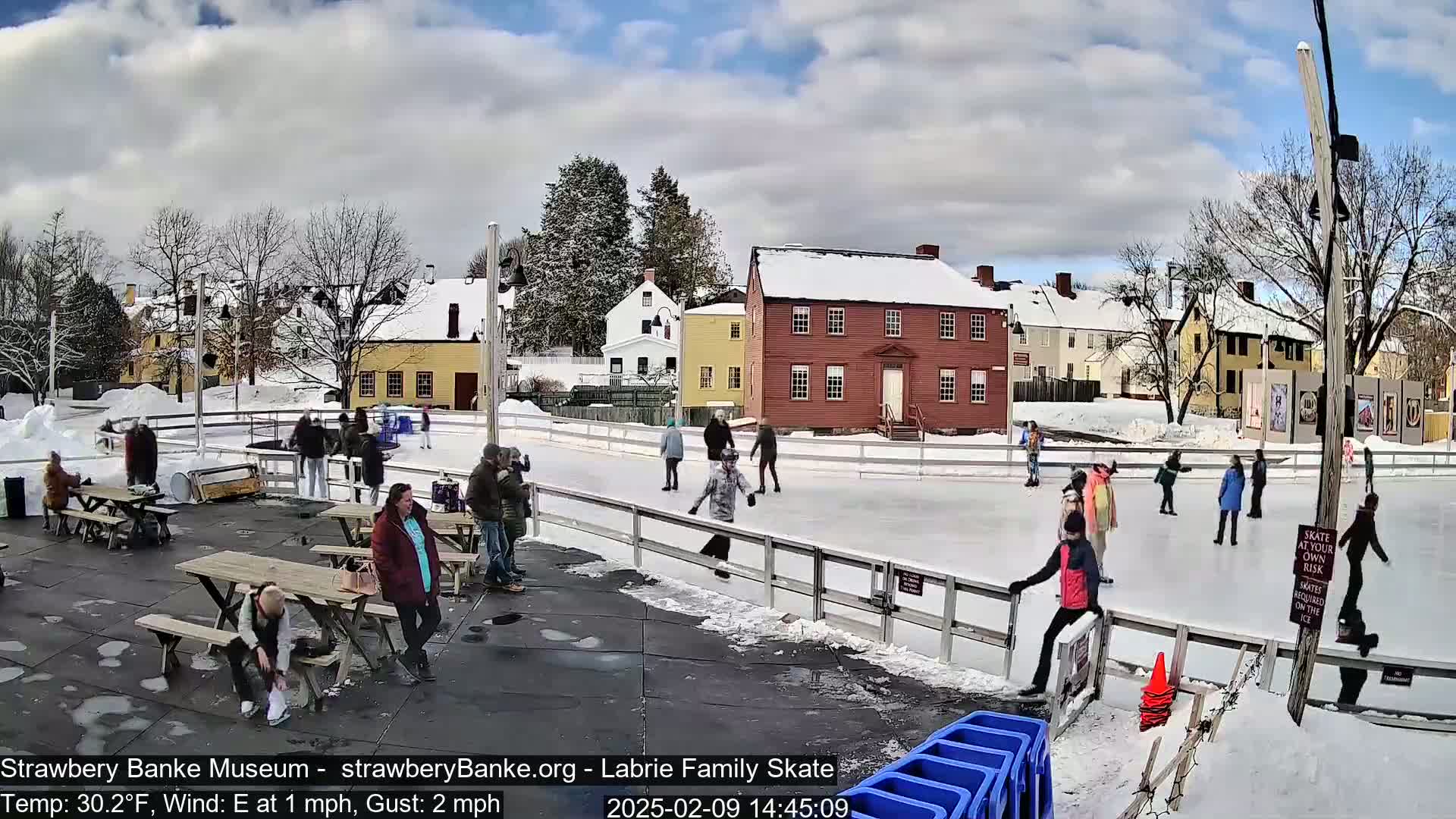 An outdoor ice rink, surrounded by snow-covered buildings and trees under a partly cloudy sky, features several people ice skating and others sitting at picnic tables nearby.