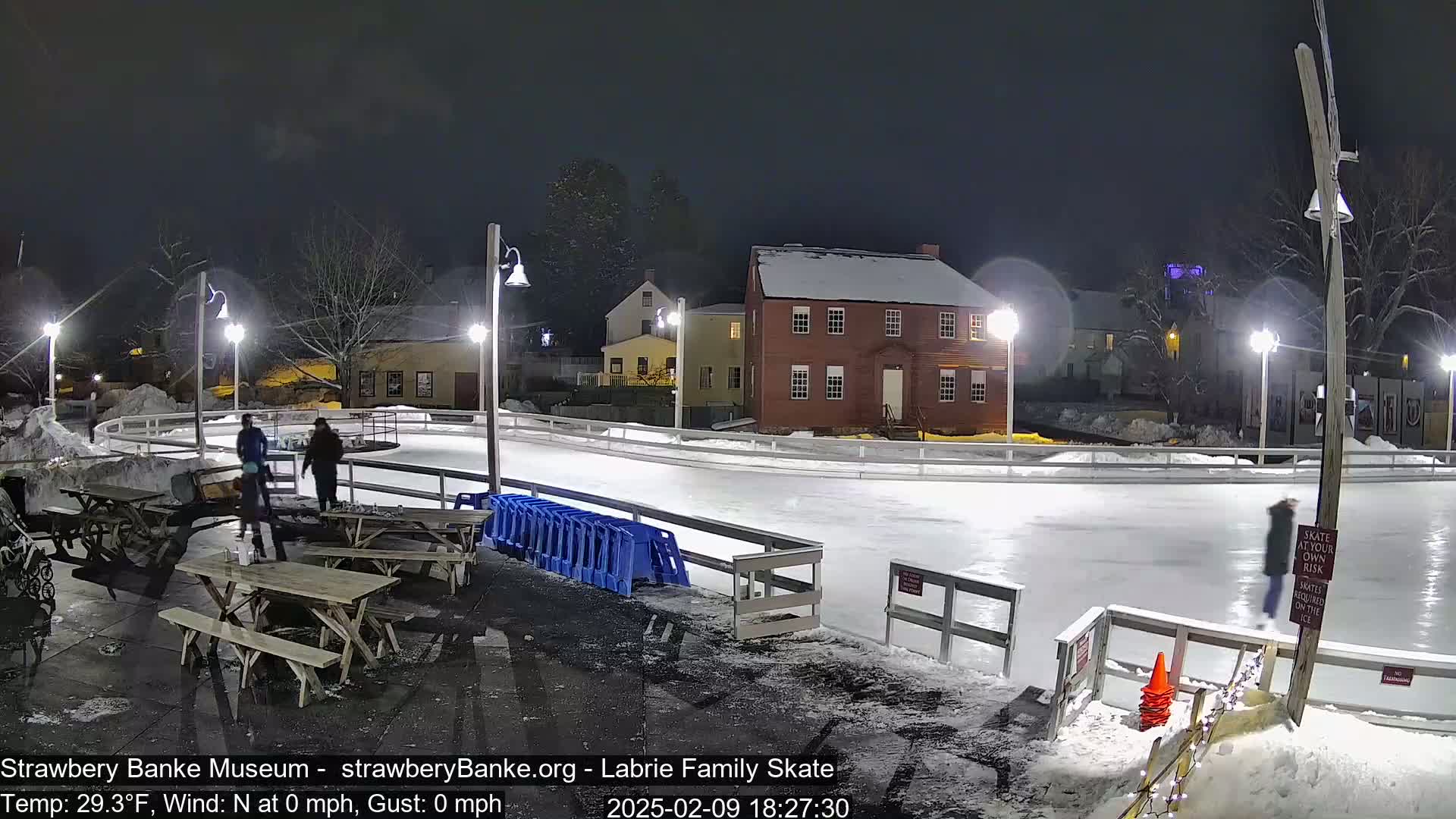 A nighttime view of an outdoor ice rink illuminated by streetlights, with a few people skating and some picnic tables nearby, next to a snow-covered building.