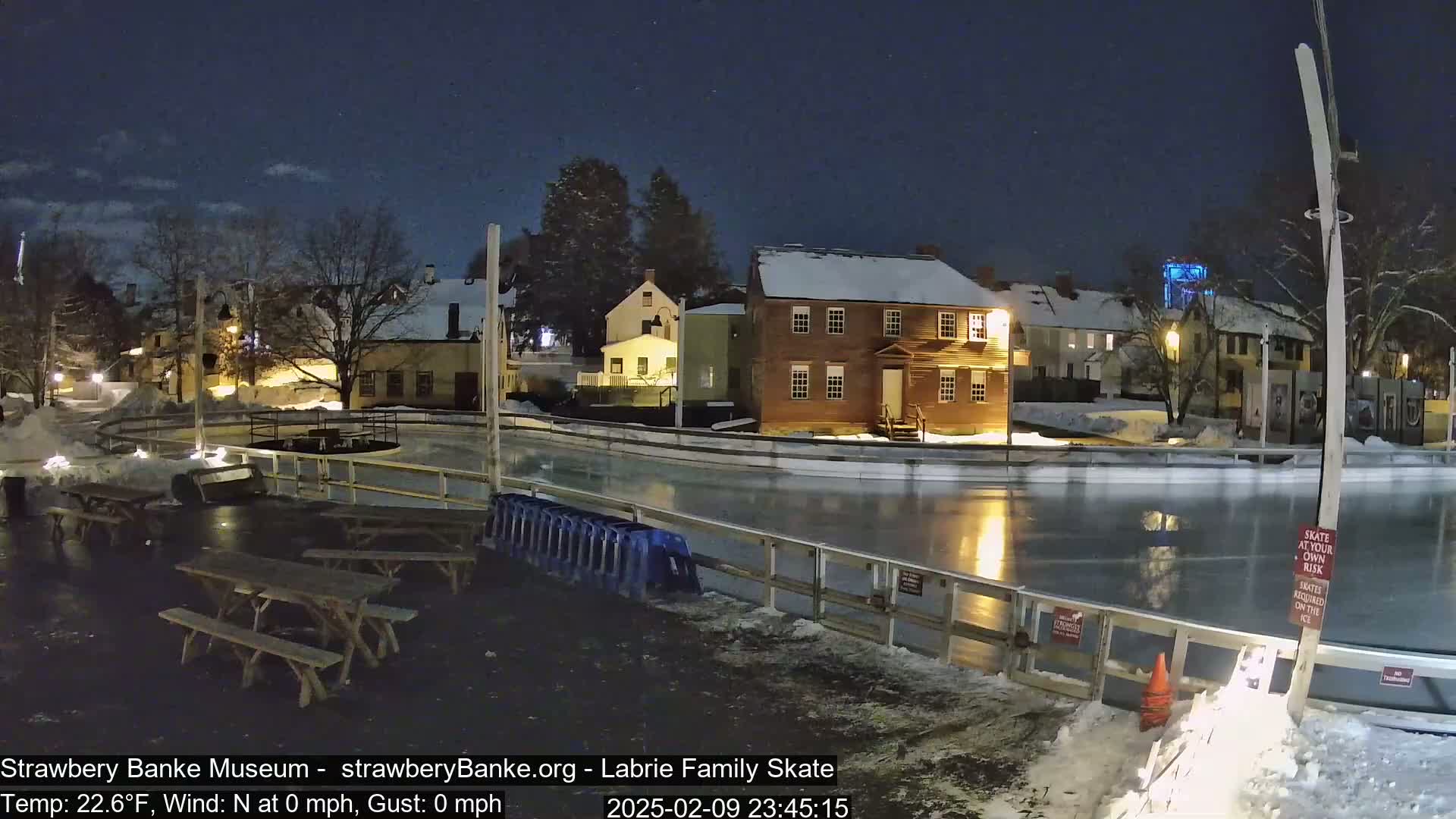 A nighttime view of an outdoor ice skating rink surrounded by snow-covered buildings under a partly cloudy sky.