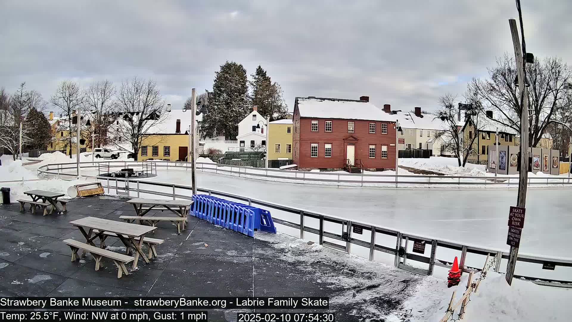A snow-covered outdoor ice skating rink is situated next to several historic buildings under a cloudy sky.