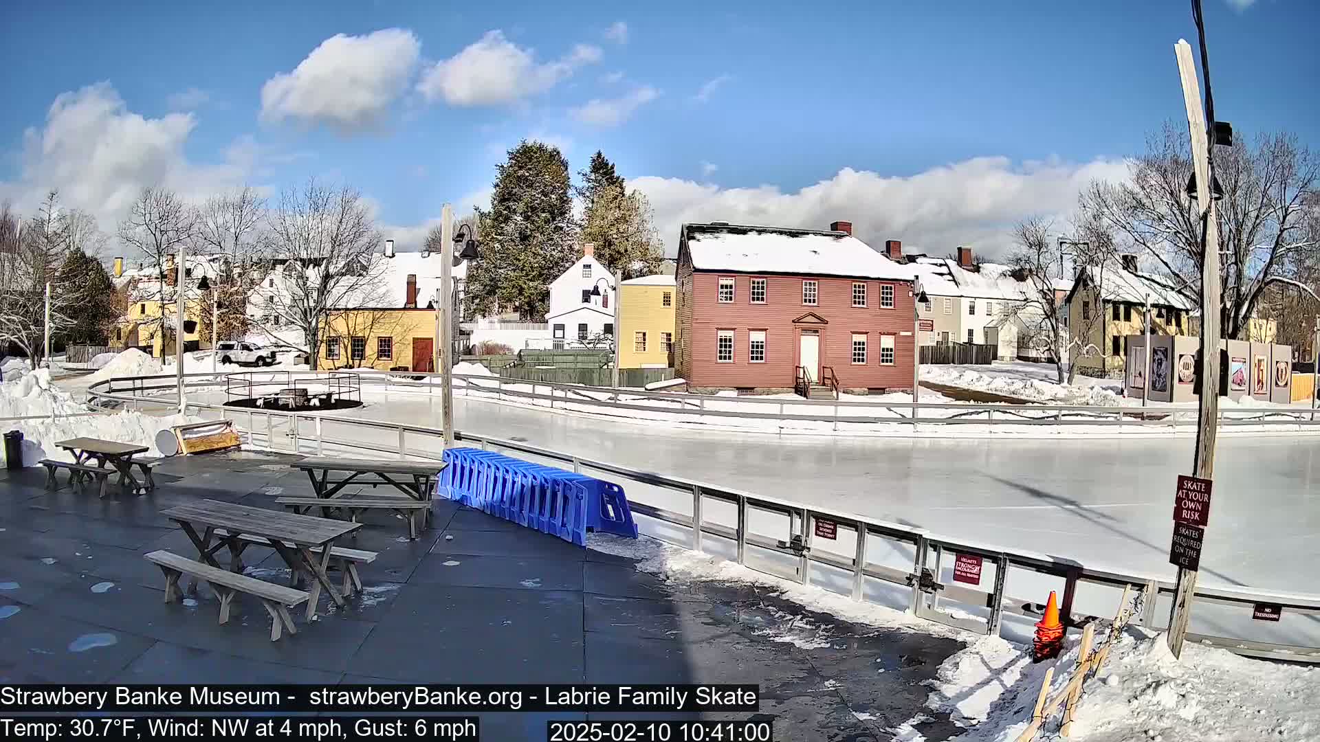 An outdoor ice skating rink is surrounded by snow-covered, historic-looking buildings under a mostly sunny sky.
