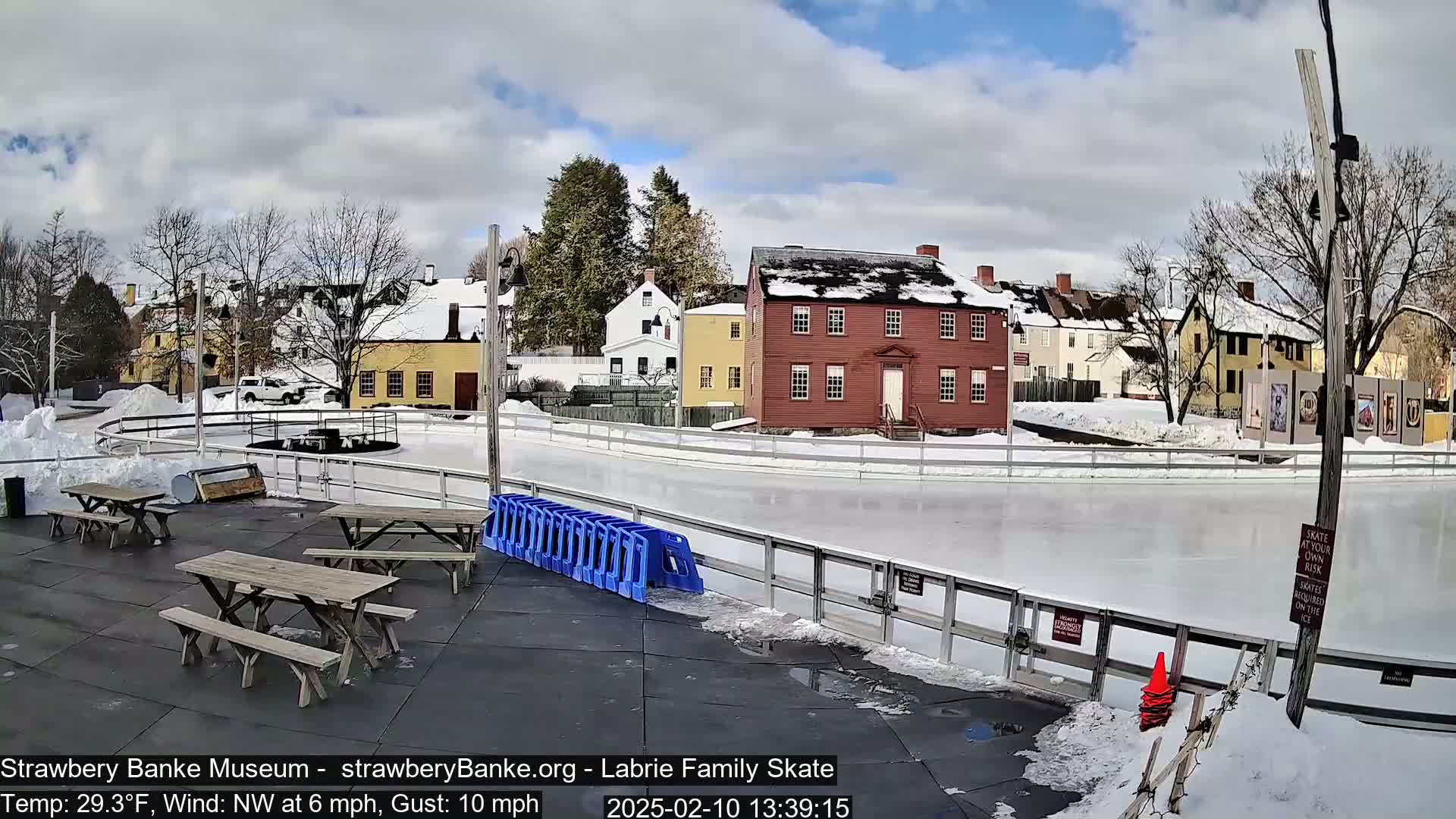 An outdoor ice skating rink, surrounded by snow-covered ground and historic-looking buildings, under a partly cloudy sky.