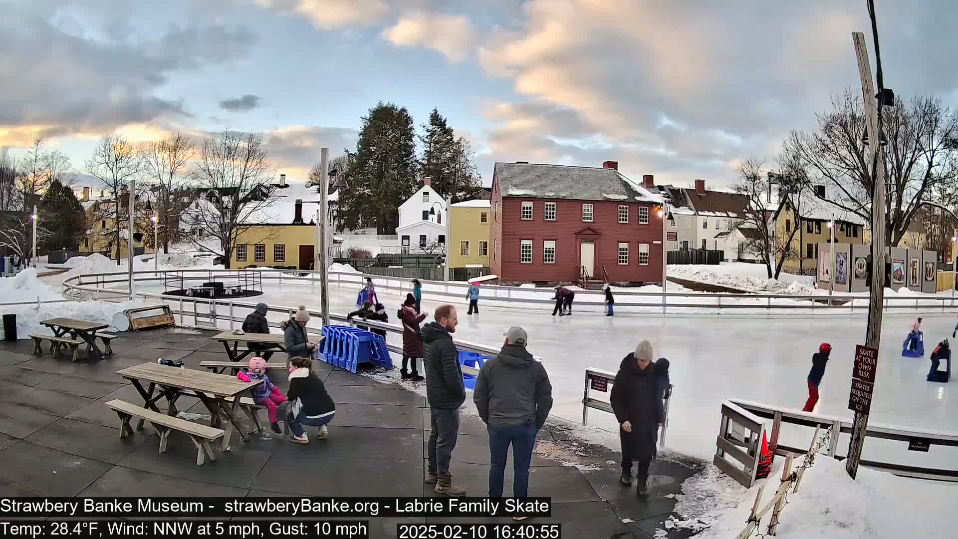 An outdoor ice skating rink, surrounded by snow-covered buildings and picnic tables, under a partly cloudy sky.