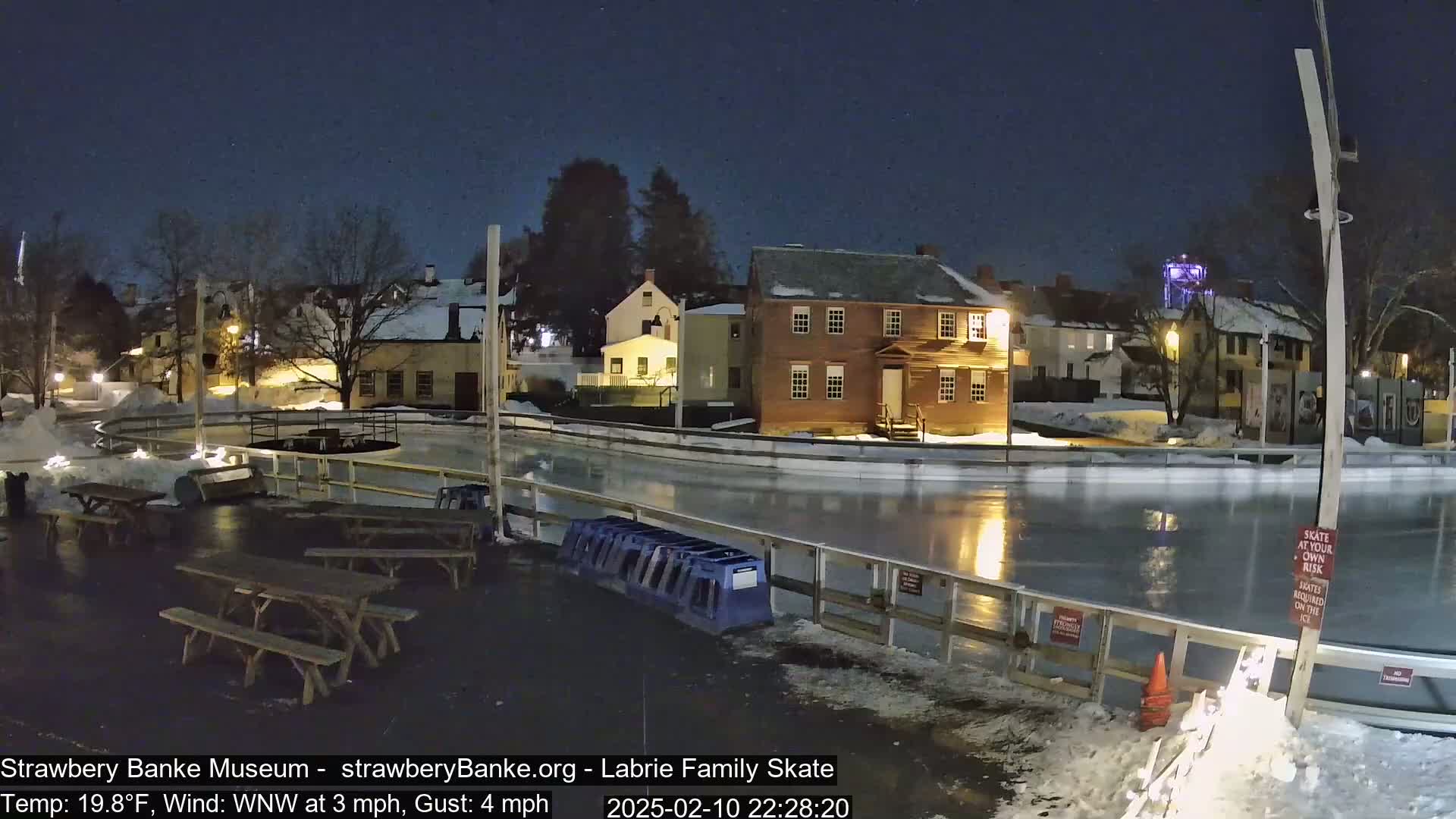 A nighttime view of an outdoor ice skating rink surrounded by snow-covered buildings and picnic tables under a clear, dark sky.