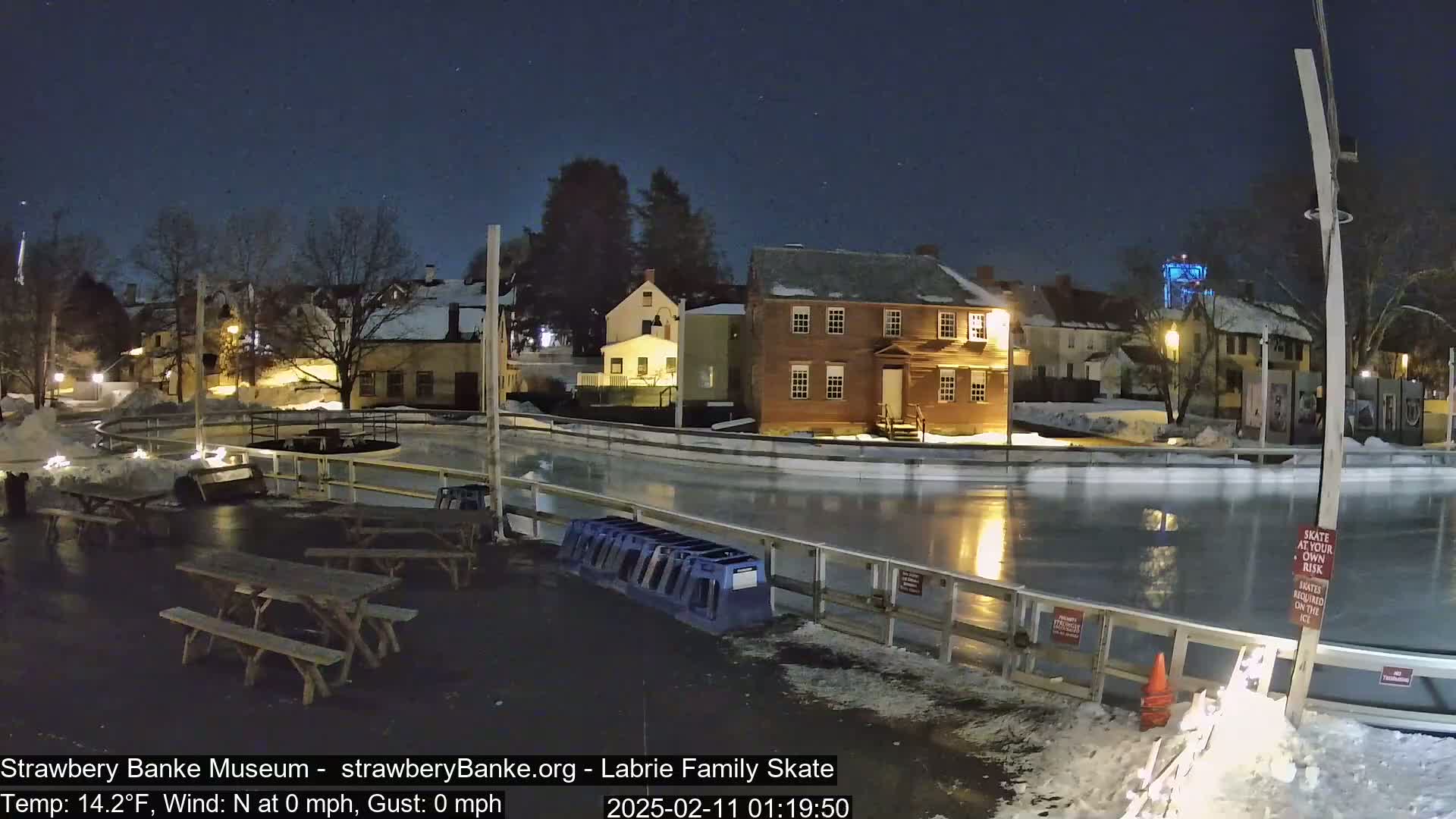 An outdoor ice skating rink at night, illuminated by lights, is situated near several historic-looking buildings under a clear, dark sky.