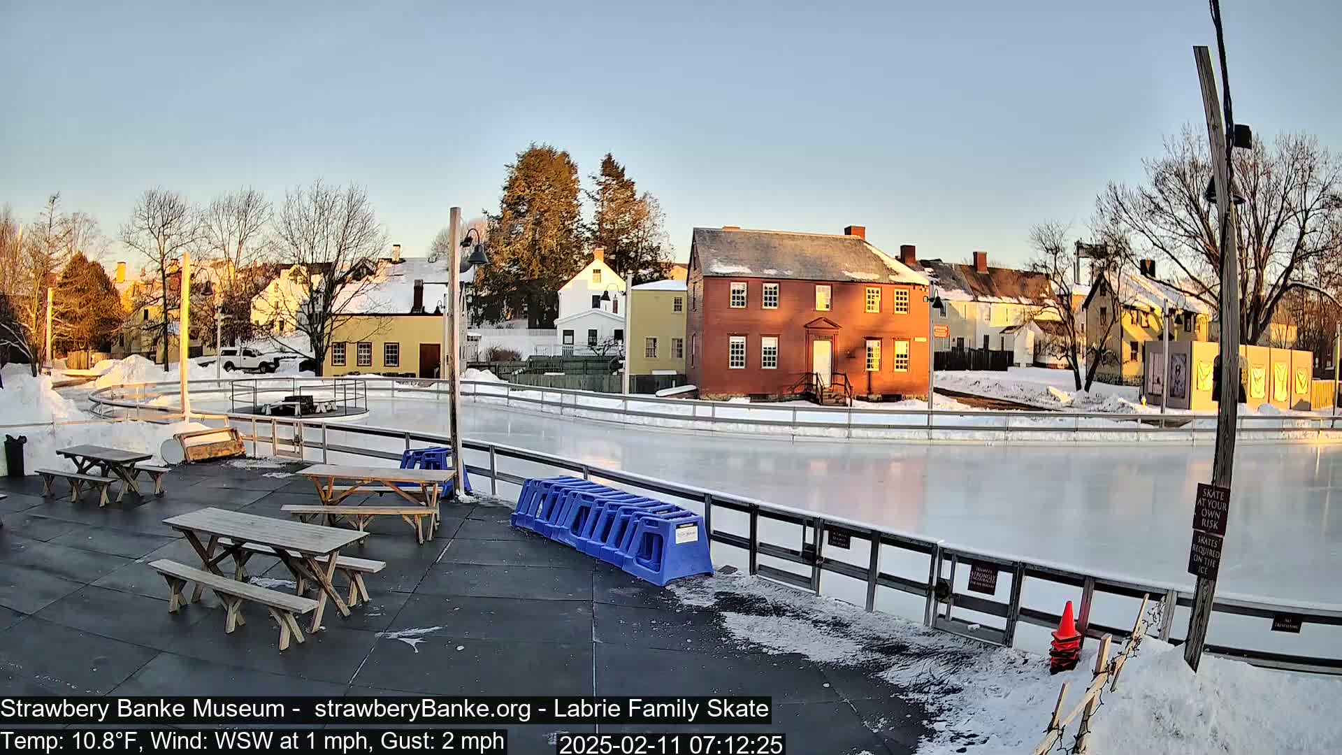 A frozen outdoor skating rink sits beside several colonial-style buildings on a sunny, cold, and slightly windy day.