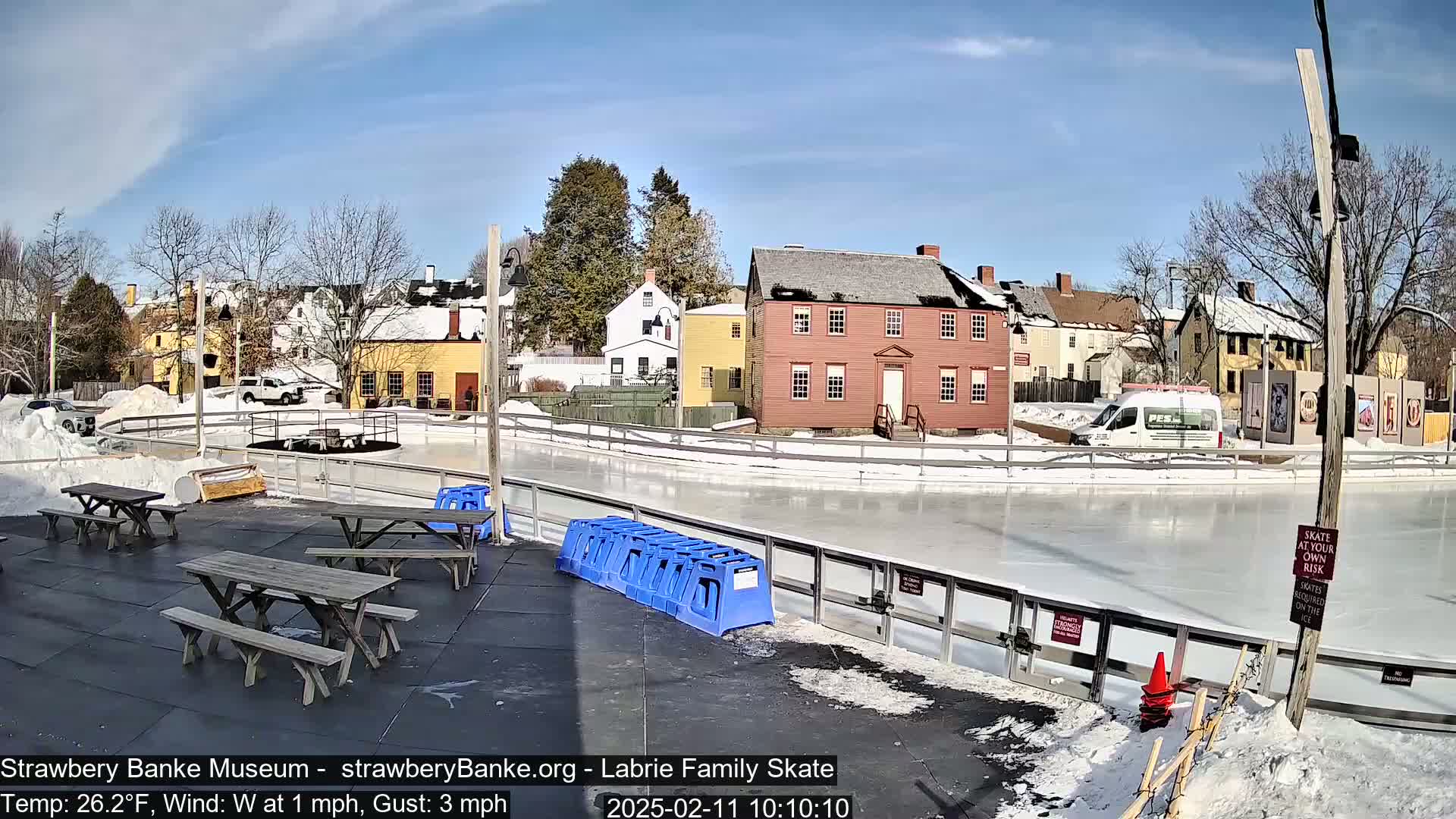 A sunny outdoor ice rink, surrounded by historic buildings and picnic tables, is partially covered in snow.