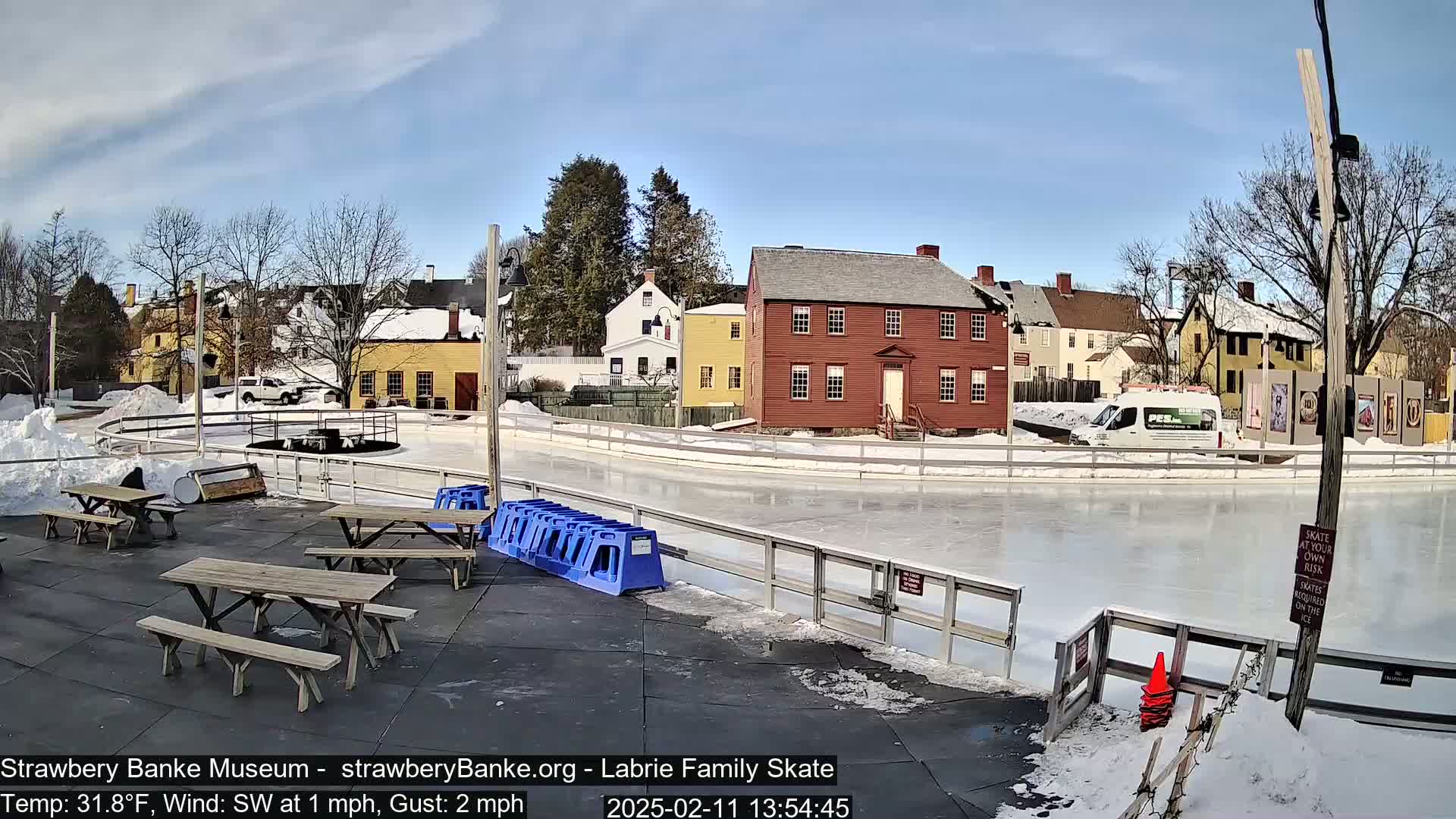 A partially frozen outdoor ice rink is situated near several historic-looking buildings on a sunny, cold day with snow on the ground.