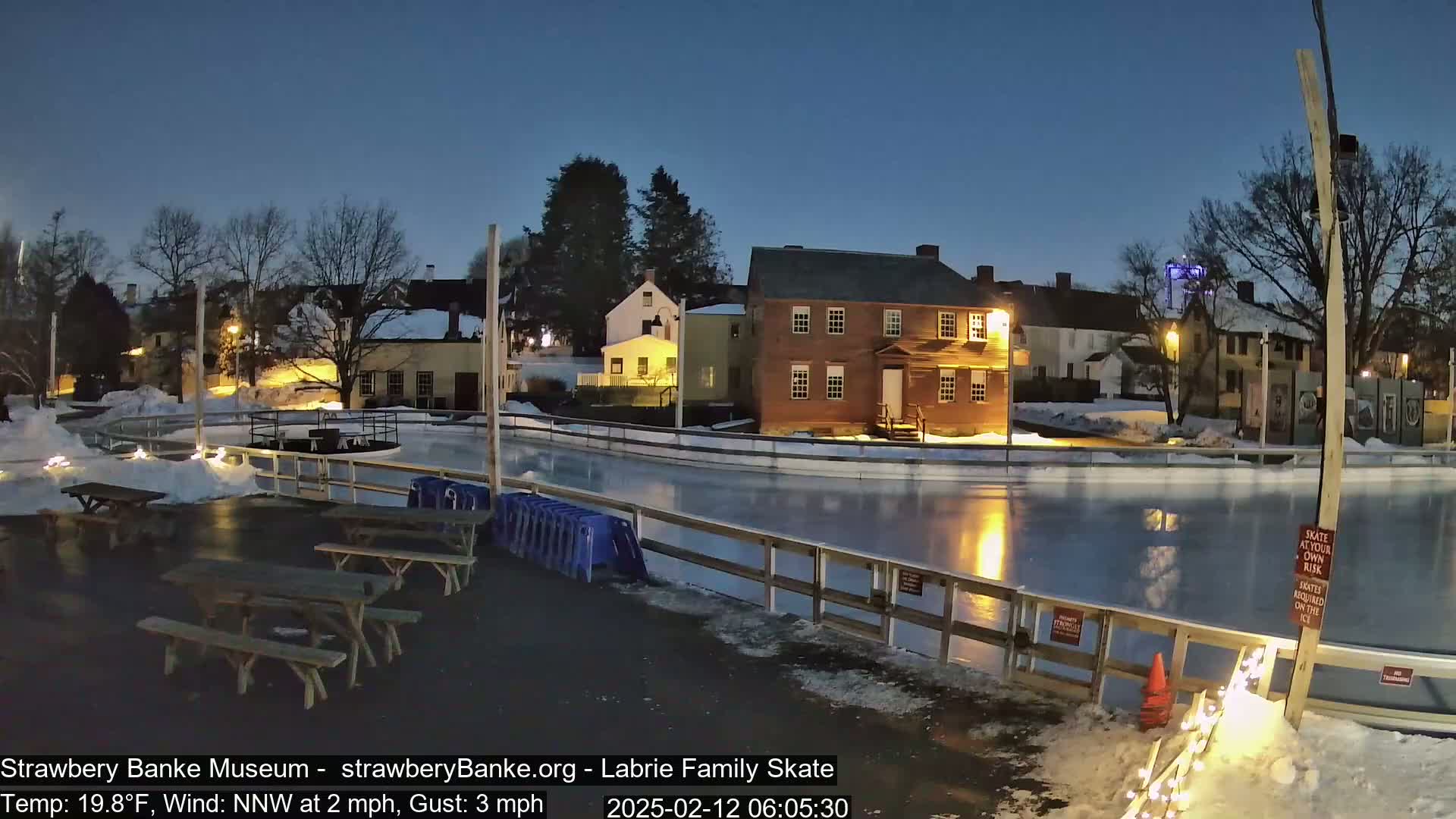 An outdoor ice rink is illuminated at night under a twilight sky, surrounded by snow-covered picnic tables and historic buildings.