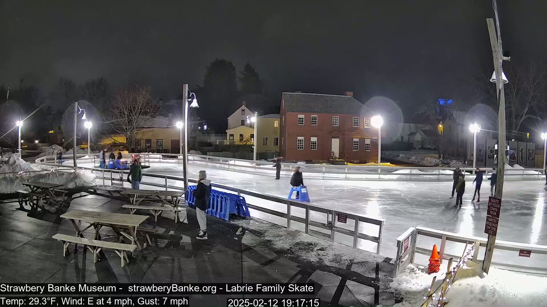At night, under a dark sky, several people ice skate on an outdoor rink illuminated by lights, surrounded by snow and picnic tables.