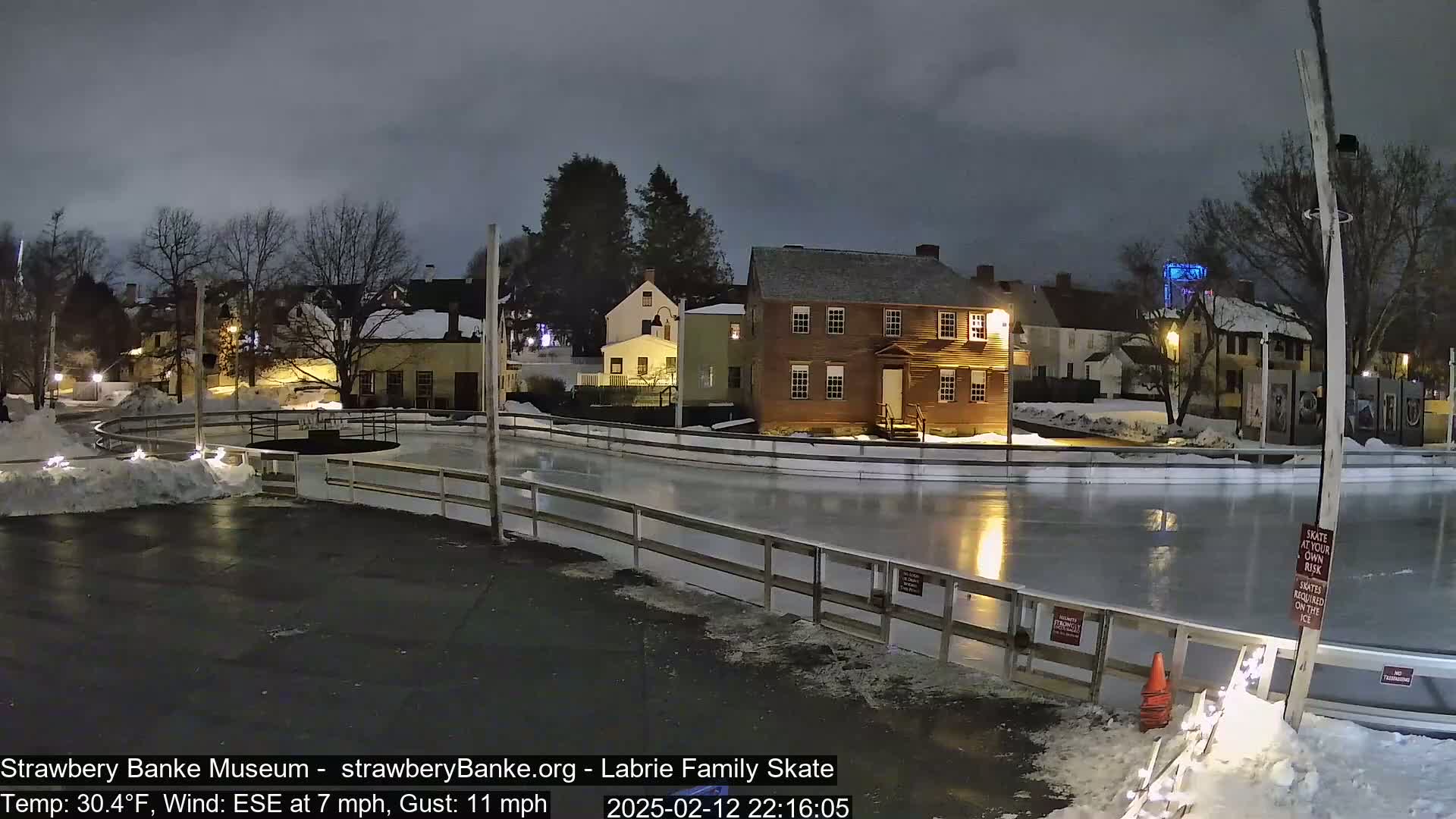 A nighttime view of an outdoor ice skating rink illuminated by lights, surrounded by snow-covered ground and historic-looking buildings under a cloudy sky.