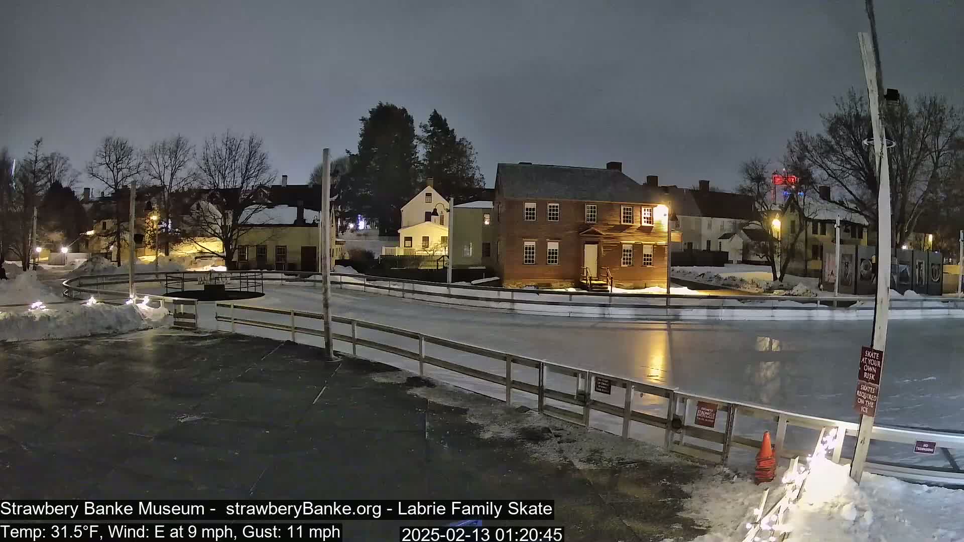 A nighttime view of an outdoor ice skating rink, surrounded by snow and historic-looking buildings, under a cloudy sky with a 9 mph east wind and gusts up to 11 mph.