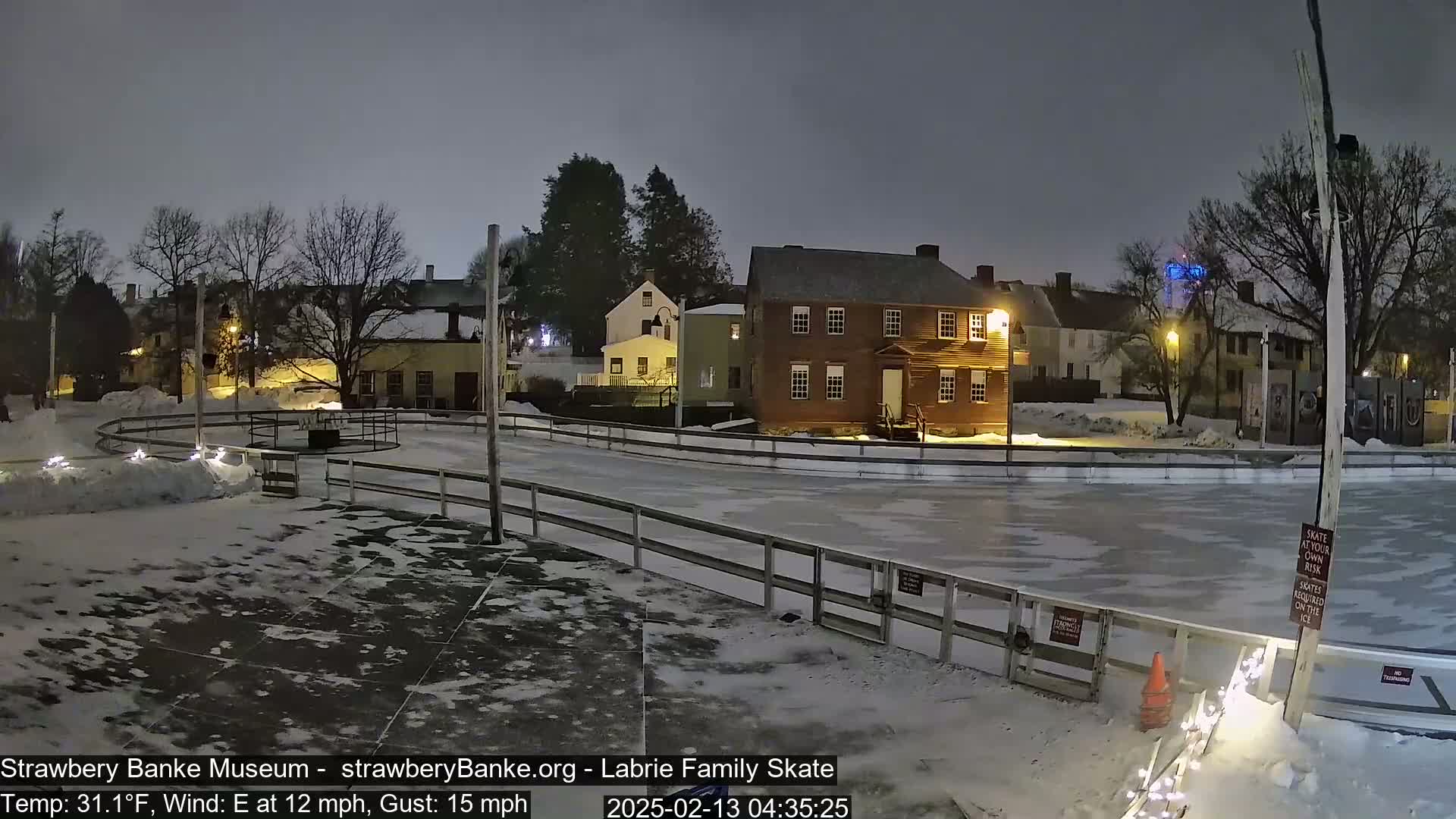 A nighttime view of an outdoor ice skating rink surrounded by snow-covered ground and historic-looking buildings under a dark, overcast sky.