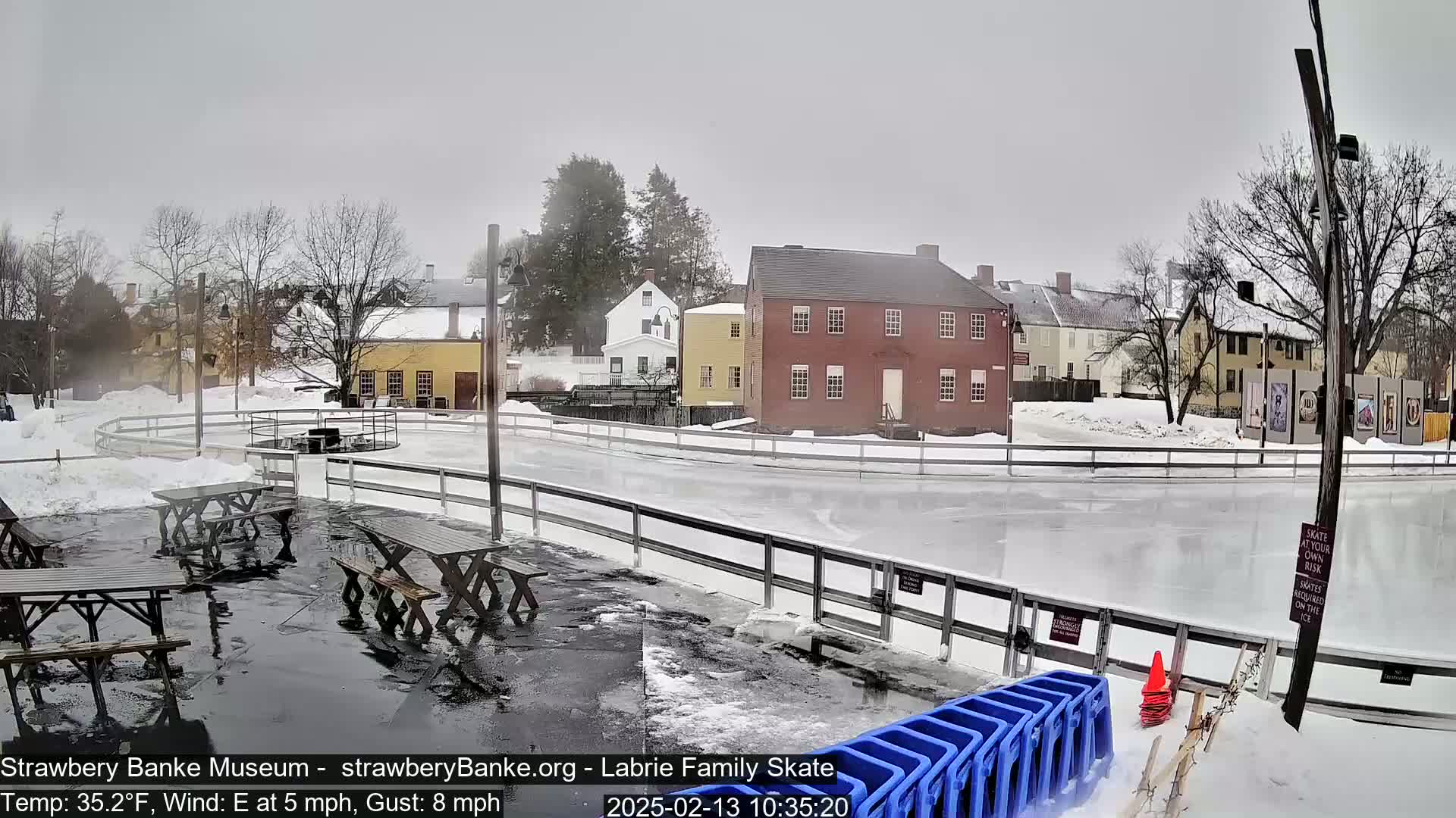 An outdoor ice skating rink, surrounded by snow-covered ground and historic buildings, under an overcast sky.