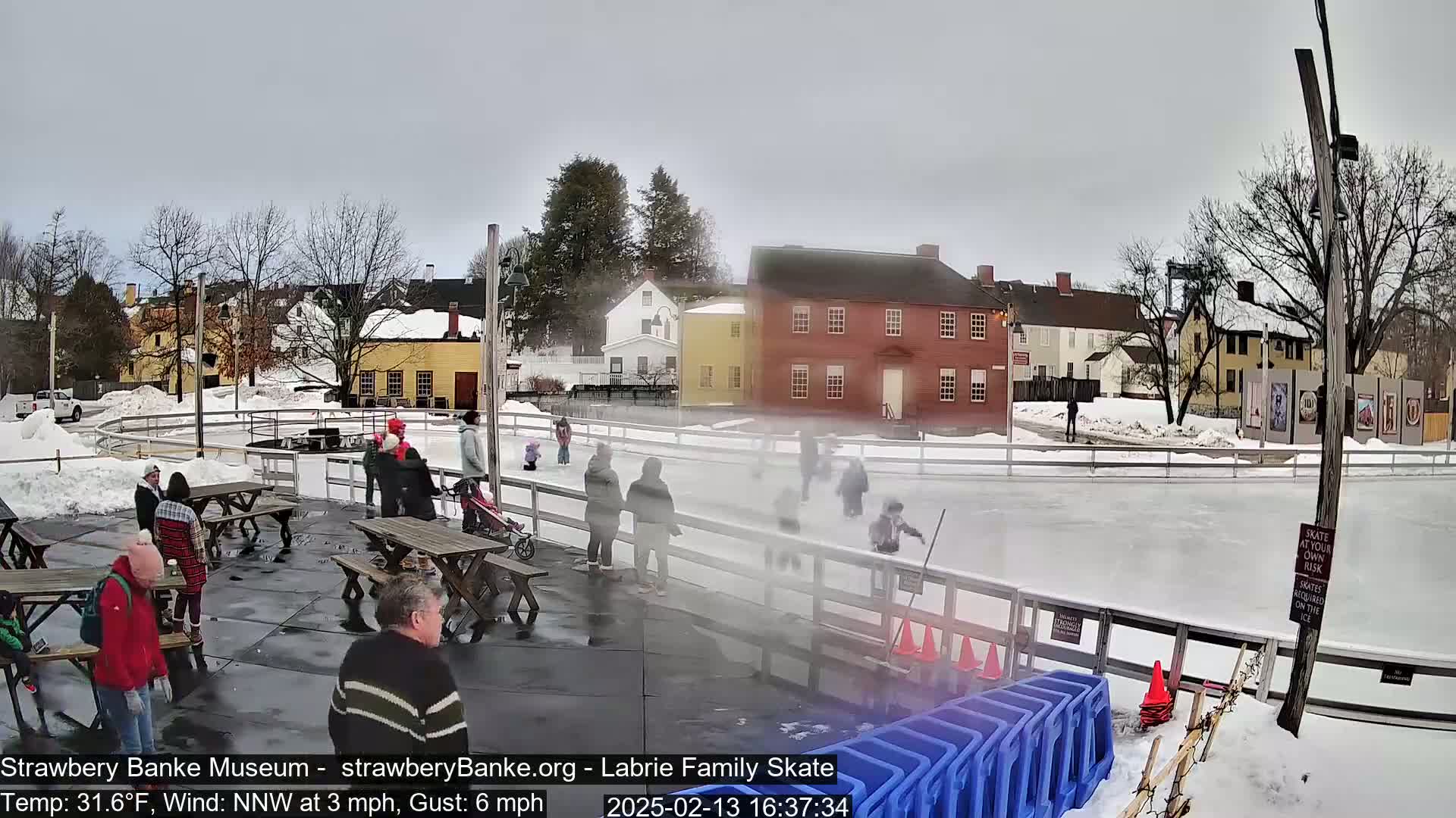 An outdoor ice skating rink with people skating and others sitting at picnic tables, surrounded by snow-covered buildings and trees under a cloudy sky.