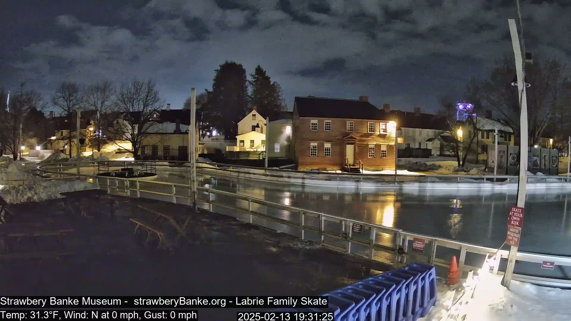 A nighttime view of an outdoor ice rink surrounded by historic buildings under a partly cloudy sky.