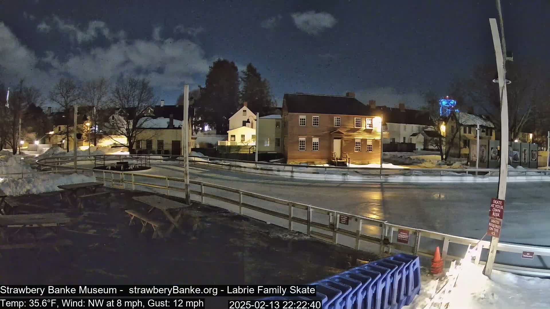 A nighttime view of an outdoor ice skating rink, illuminated by lights, surrounded by snow-covered ground and historic buildings under a partly cloudy sky.