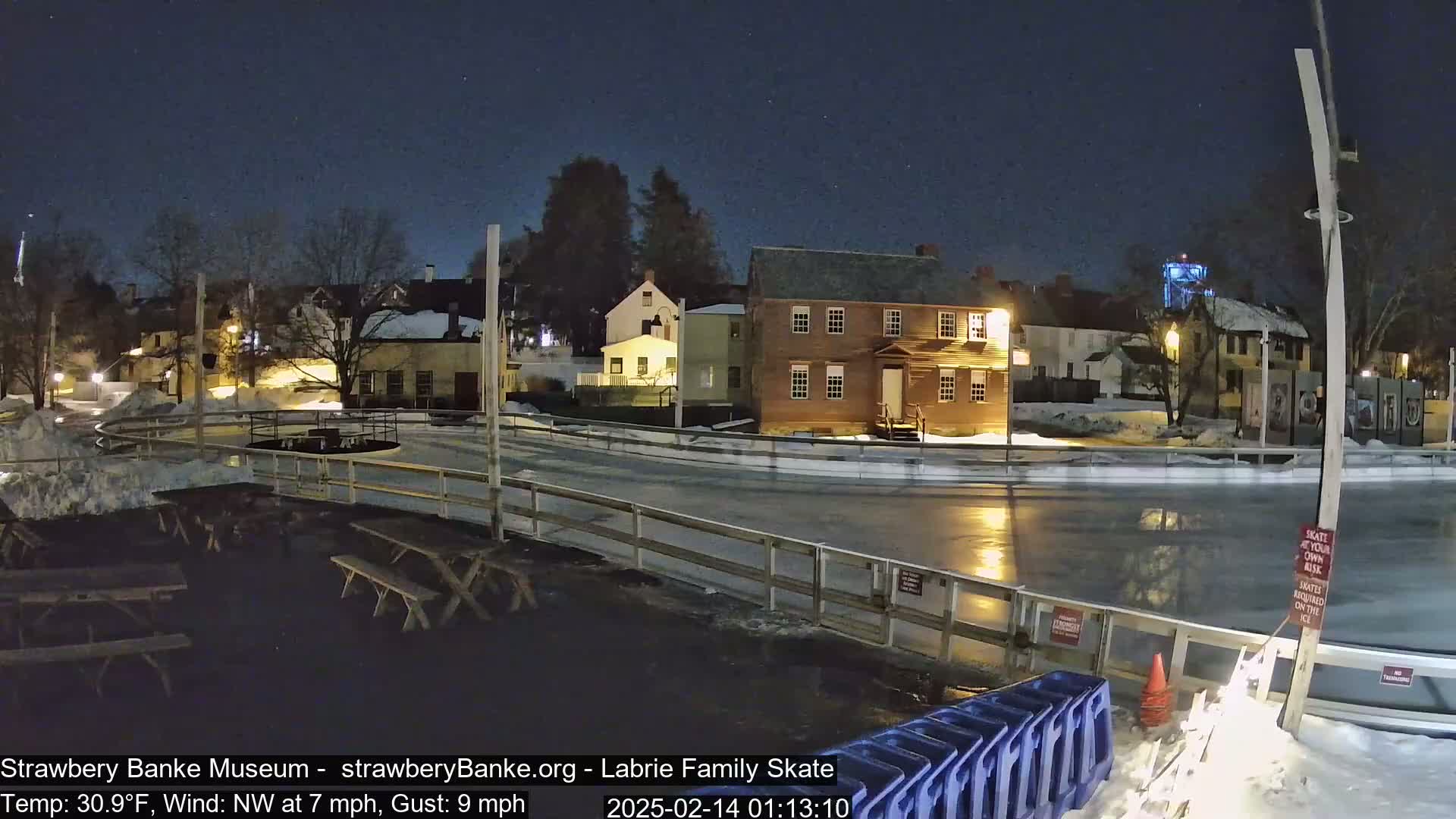 A nighttime view of an outdoor ice rink surrounded by historic-looking buildings under a clear, starry sky, with snow on the ground and a northwest wind.