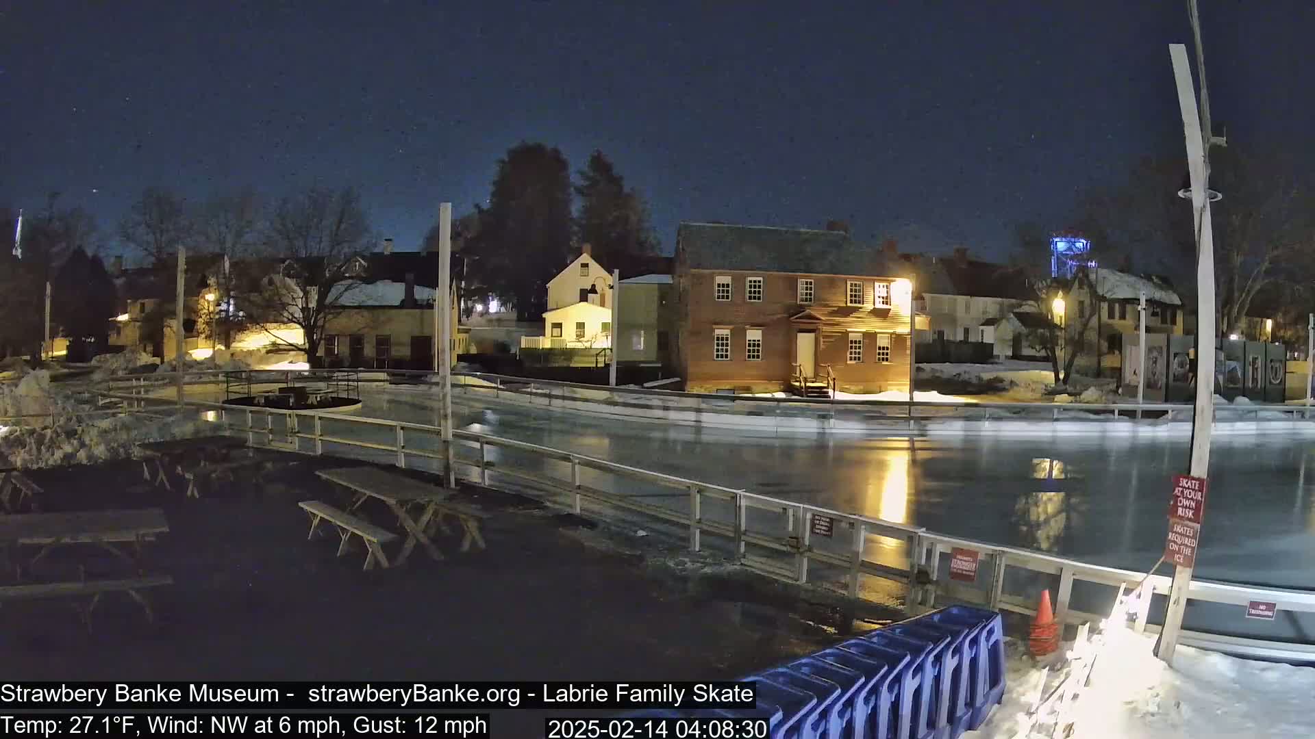 An outdoor ice skating rink is illuminated at night under a clear, starry sky, surrounded by historic buildings and snow-covered ground.