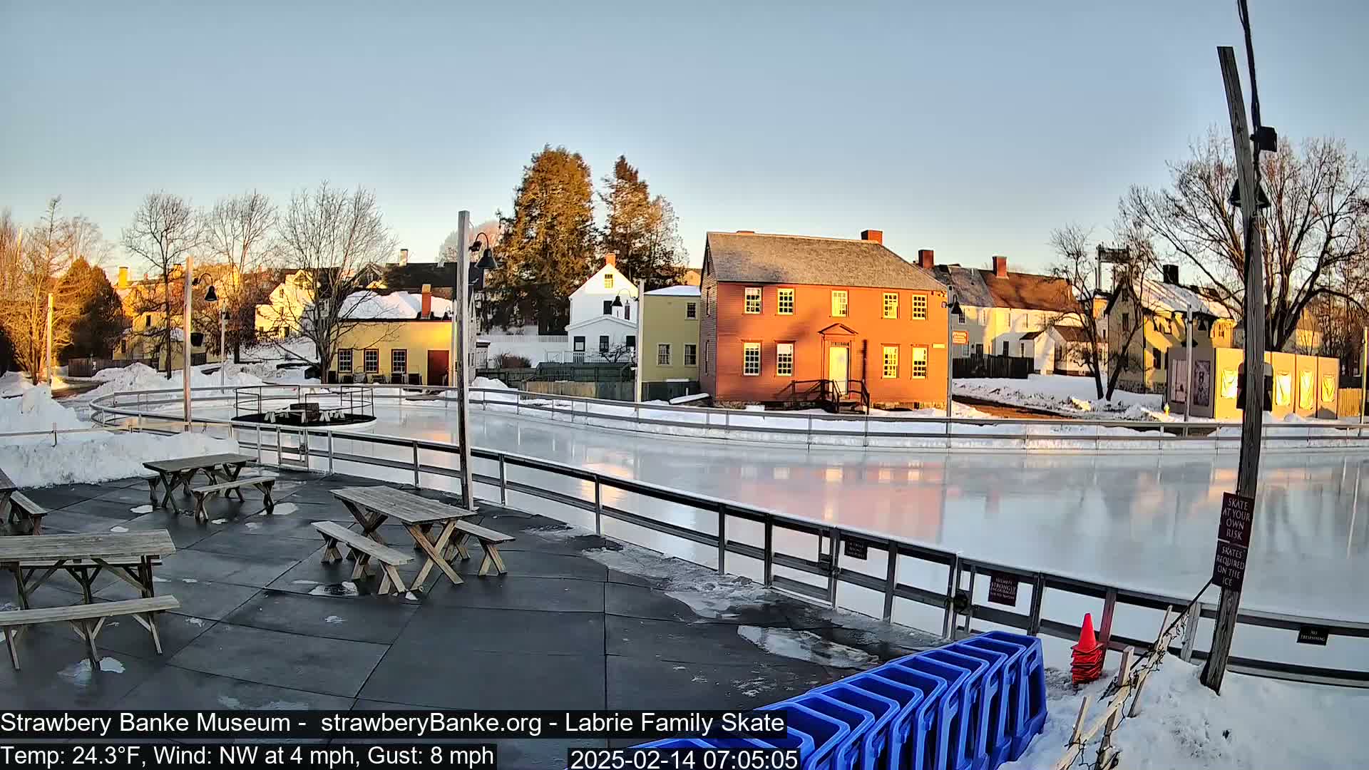 An outdoor ice skating rink, surrounded by picnic tables and historical buildings, is bathed in the soft light of sunrise on a cold, partly sunny morning with a northwest wind.